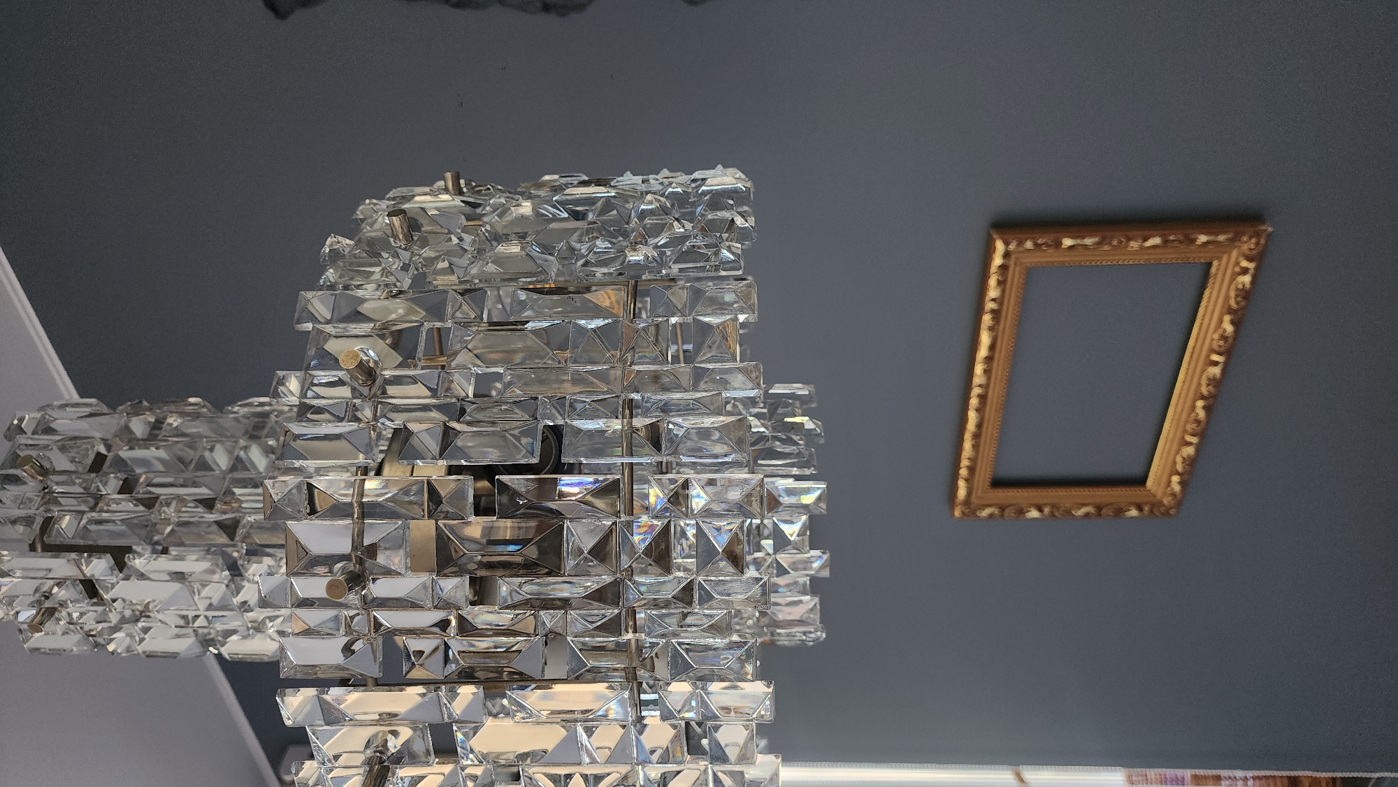 The image shows a portion of a ceiling, likely indoors. A large, multifaceted crystal chandelier dominates the left side of the frame. To the right is a tilted, ornate gold frame hanging on a wall or ceiling. The background is a flat, dark gray surface. There is no indication of the location being Smolensk, Russia, or any other identifiable location. There are no people visible in the image, and there are no identifiable actions or interactions taking place.