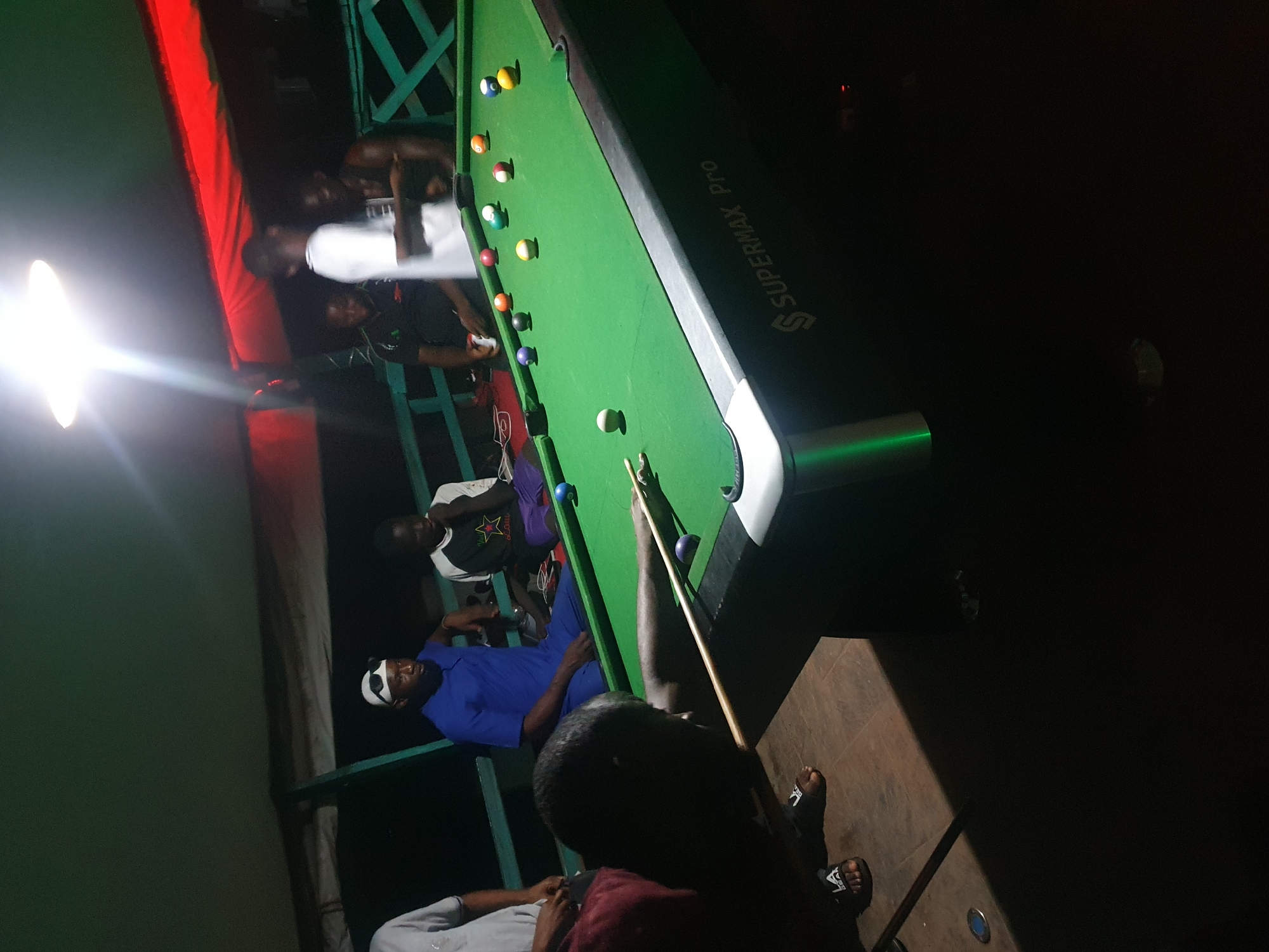 In Lokoja, Nigeria, a group of men are engaged in a game of pool. The scene is set in an outdoor or semi-outdoor area, possibly a bar or recreational spot, with a green pool table as the central focus. A bright overhead light illuminates the table and some of the surrounding area, casting strong shadows.
Several individuals are visible around the pool table. One man, wearing a blue dashiki and white sunglasses, is seated. Another man, wearing a black t-shirt with a graphic, is also seated. A third man, in a white t-shirt, is standing and appears to be interacting with others. In the foreground, a man is in a crouched position, aiming to strike a billiard ball with a cue stick. He is wearing a maroon shirt and dark sandals. Another man, partially visible on the left, wears a grey t-shirt.
The pool table itself is branded with "SUPERMAX Pro". Various billiard balls, including solid colors and striped balls, are scattered across the green felt surface. The background suggests a rustic setting with wooden structures and possibly a covered awning overhead, indicated by the red fabric visible. The overall ambiance appears casual and social.