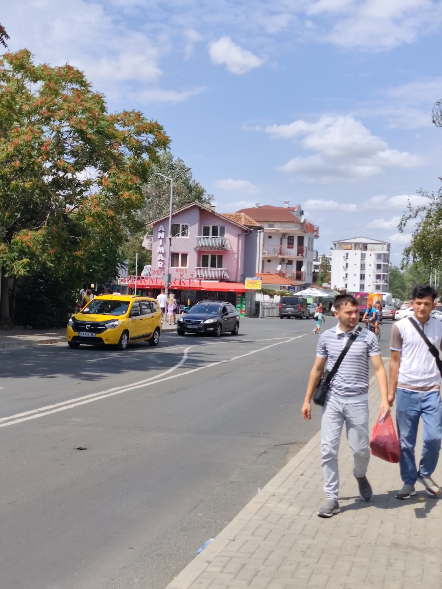 The image depicts a street scene in the city of Nesebar, Bulgaria. On the left, a yellow Dacia Lodgy taxi is driving on the road. To the right of the taxi, a black car is also on the road. A pink building with balconies and "ANMAR" signage is visible behind them, with a red awning extending from its ground floor. Further in the background, a modern white building stands. Two young men are walking on the sidewalk in the foreground, one carrying a black bag over his shoulder and the other holding a red shopping bag. The sky is blue with scattered white clouds, and trees with red blossoms are present on the left side of the image. The overall environment suggests a sunny day in a commercial or residential area.