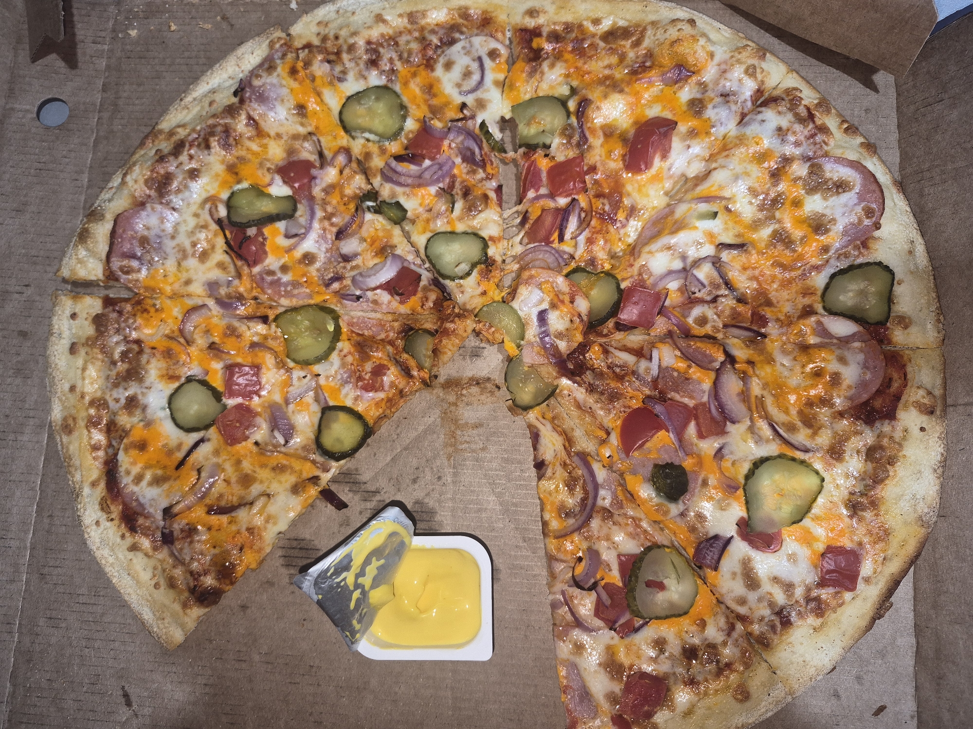 The image shows a pizza in a cardboard box. The pizza is sliced and has toppings including pickles, tomatoes, red onions, and cheese. A small container of yellow sauce is placed next to a slice of the pizza. The pizza appears to be in a box on a brown surface, possibly a table or the floor of a room. There are no people visible in the image. The image does not provide any information about the location of the city of Chaikovsky, Russia.