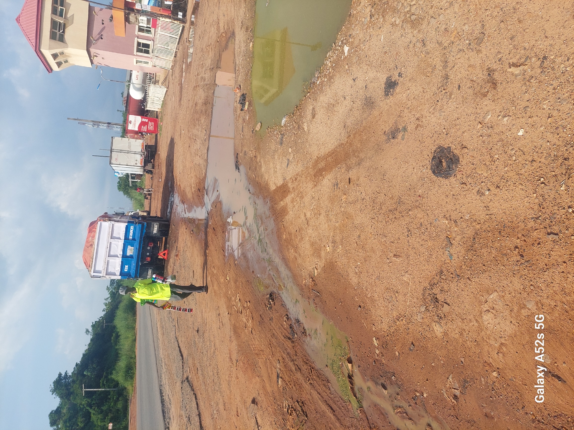 In Awkuzu, Nigeria, a man in a bright yellow shirt and grey pants walks across a dirt road littered with debris and puddles of muddy water. He carries a bundle of what appear to be small, rectangular items, possibly packaged goods, in his left arm, and a green spray can and a plastic bag in his right hand. Behind him, a large blue and white truck with a covered bed is parked on the side of the road, its hazard lights on. A red triangular warning sign is placed behind the truck. In the background, several other vehicles are visible, including a white truck and a tanker. To the right, a distinctive pink and tan tower-like structure with windows and a red roof stands near a white fence. The sky above is partly cloudy.