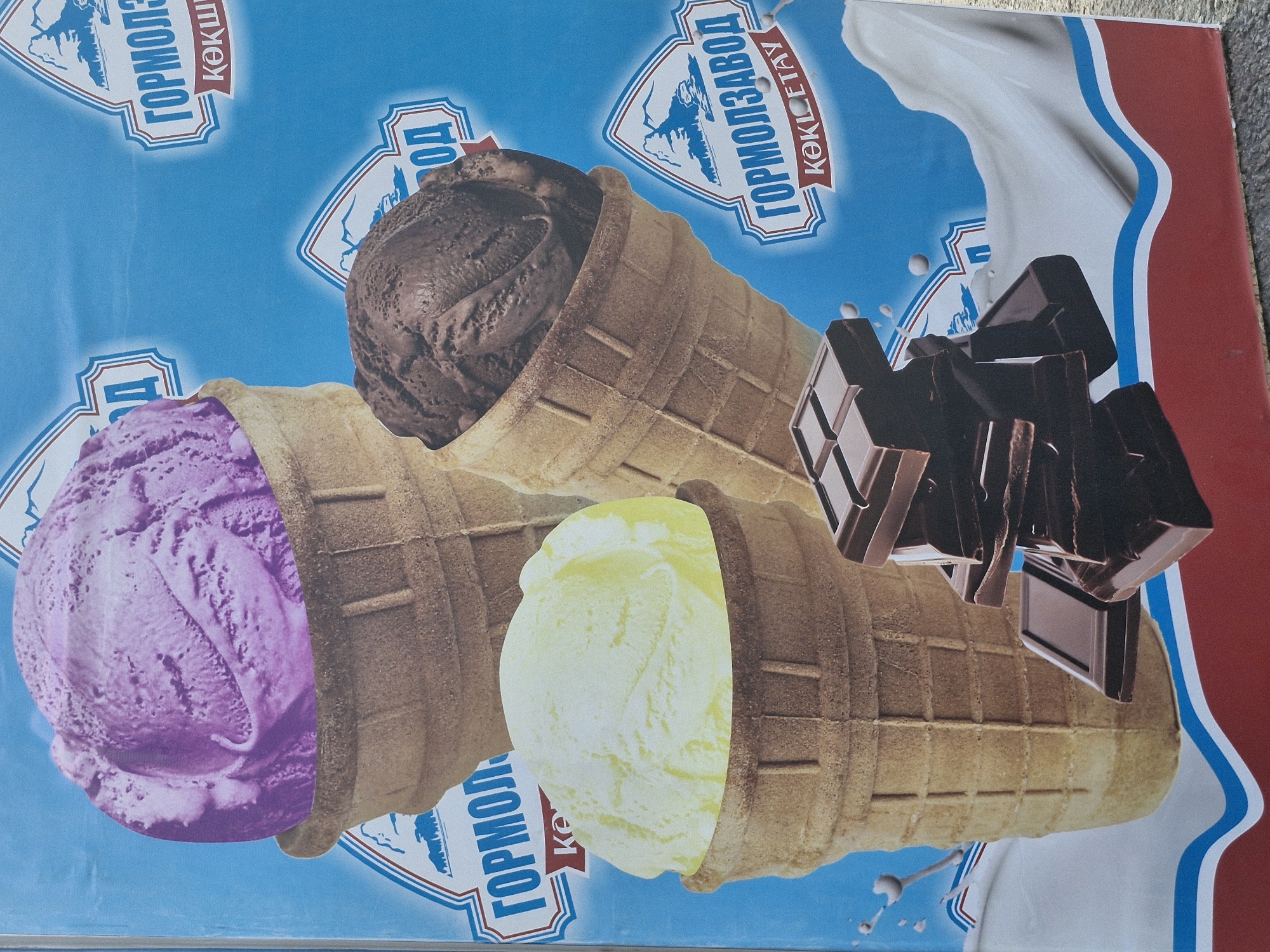 The image is a close-up of an advertisement for ice cream, featuring three ice cream cones and pieces of chocolate. The background is a bright blue with some red and white graphic elements, likely from the advertisement's design.
In the foreground, to the right, is a stack of dark chocolate bars. To the left and slightly behind the chocolate, a waffle cone holds a scoop of light yellow ice cream. Further to the left and higher up, another waffle cone holds a large scoop of purple ice cream. Behind and to the right of the yellow ice cream cone, a third waffle cone holds a scoop of dark brown, likely chocolate, ice cream.
Several emblems or logos are visible in the background. One, on the left, appears to have blue text reading "ГОРМОЛЗАВОД" (Gormolzavod) above red text that is partially obscured. Another, on the right, also features "ГОРМОЛЗАВОД" in blue text within a white, shield-like shape with red borders, above red text that reads "КӨКШЕТАУ" (Kokshetau), indicating the location in Kokshetau, Kazakhstan. Smaller versions of these logos are also present. Splashes of white liquid, presumably milk or cream, are depicted around the ice cream and chocolate. The overall scene is a vibrant and appealing depiction of frozen treats.