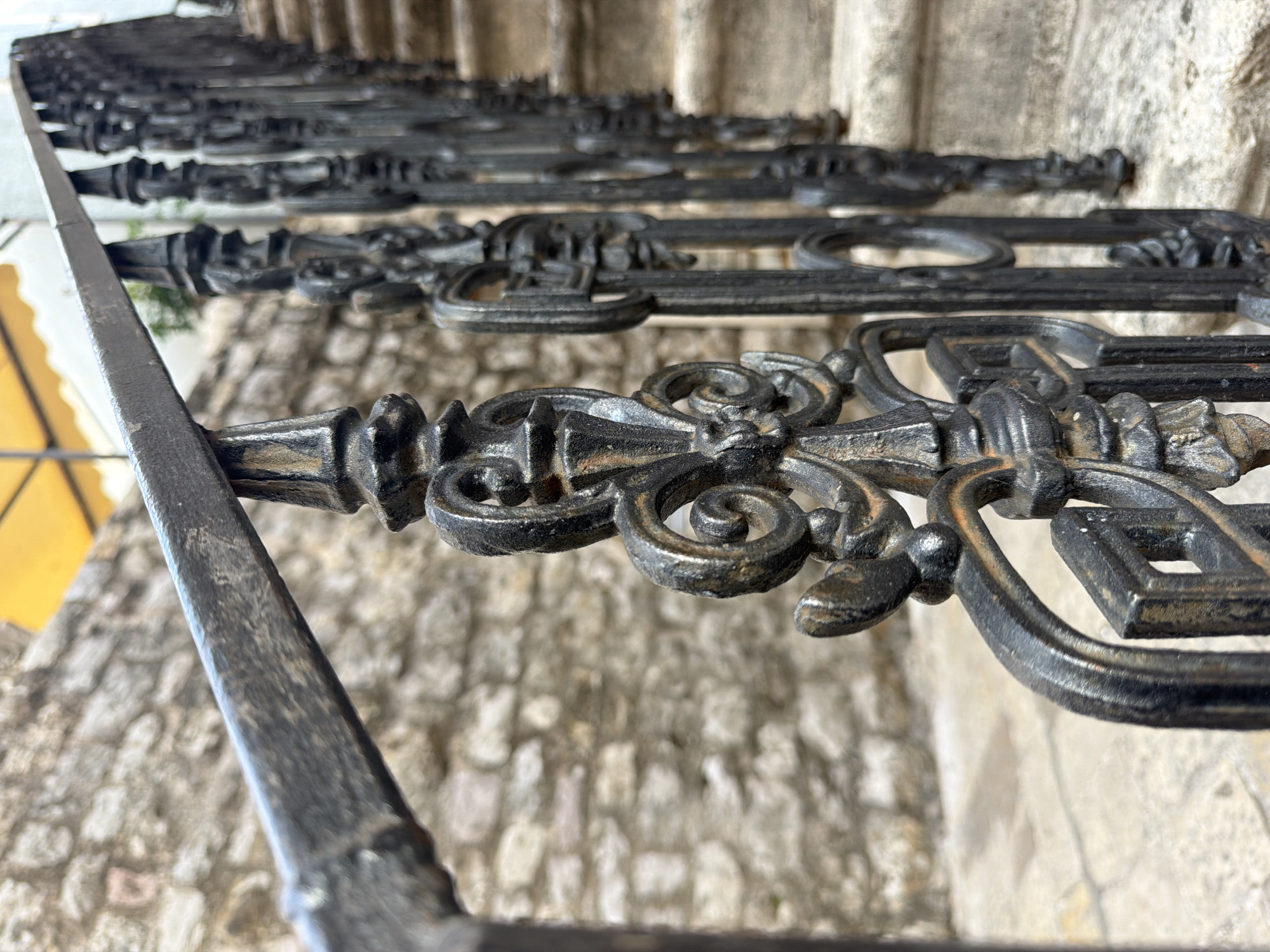 A close-up, slightly skewed perspective reveals an ornate, dark metal railing running along a stone staircase in Budva, Montenegro. The focus is sharpest on the foreground railing section, showcasing intricate, repeating designs that feature stylized floral elements and geometric shapes. The railing is patinated with rust, adding textural contrast.
The background consists of rough-hewn, light-colored stone steps and wall, which are slightly blurred due to the shallow depth of field. A hint of yellow can be seen on a building to the left of the frame.