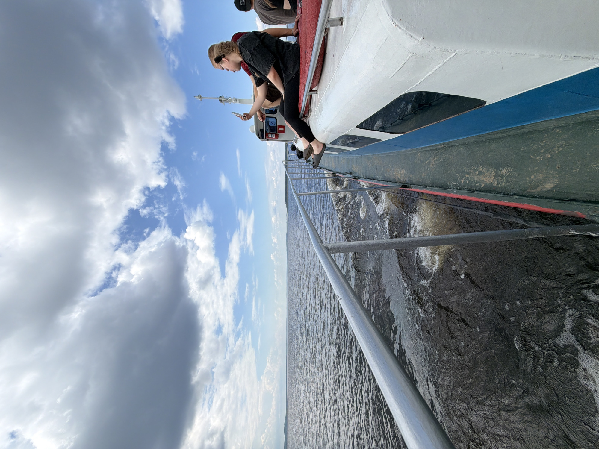 The image shows a view from the side of a boat traveling on a body of water under a cloudy sky. A young woman with braided hair, wearing a black t-shirt and black leggings, is sitting on the edge of the boat's deck, holding a smartphone and taking a selfie. She is wearing flip-flops. Another person, wearing a dark shirt, is visible behind her. The boat has a white upper structure with blue trim along the side. The water is dark and choppy, with white foam trailing behind the boat. In the distance, a low-lying shoreline is visible. The sky is filled with a mix of dark and light gray clouds, with patches of blue sky peeking through. The scene is a candid shot of people enjoying a boat trip.
