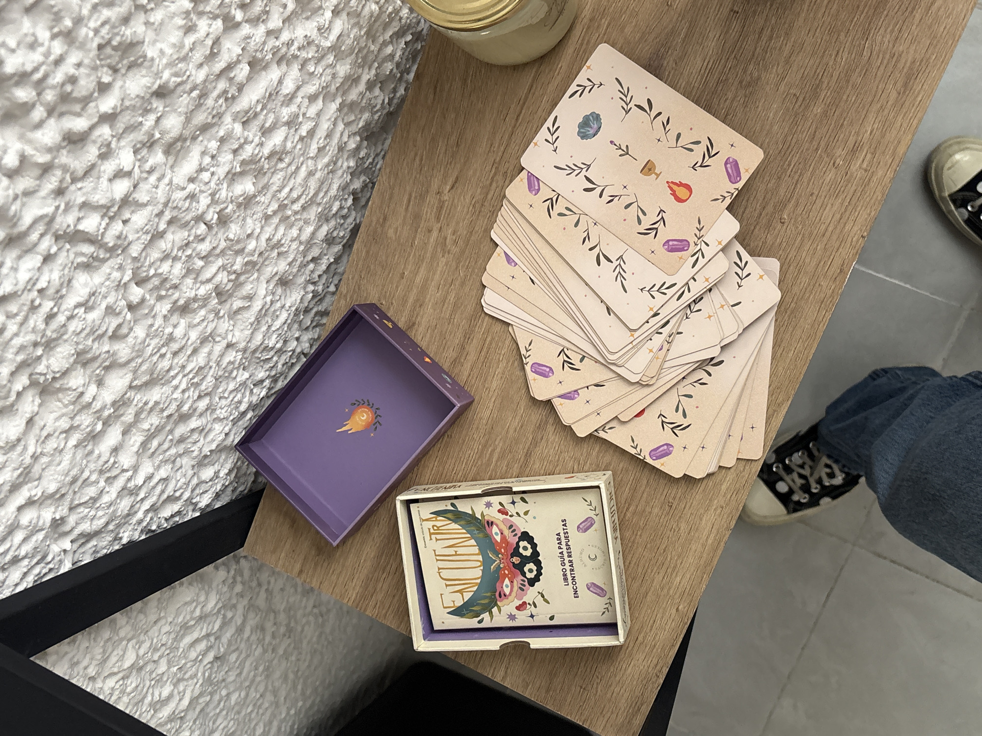 The image shows a tabletop scene with tarot cards, a purple box with a gold emblem, and a guidebook in a box with the Spanish word "ENCUENTRA." The tarot cards are spread out next to the box, which appears to be their storage container. Part of a person wearing jeans and black-and-white patterned sneakers is visible in the bottom right corner. The tabletop is light brown wood. A white, textured wall is in the background. The location is Puebla, Mexico.
