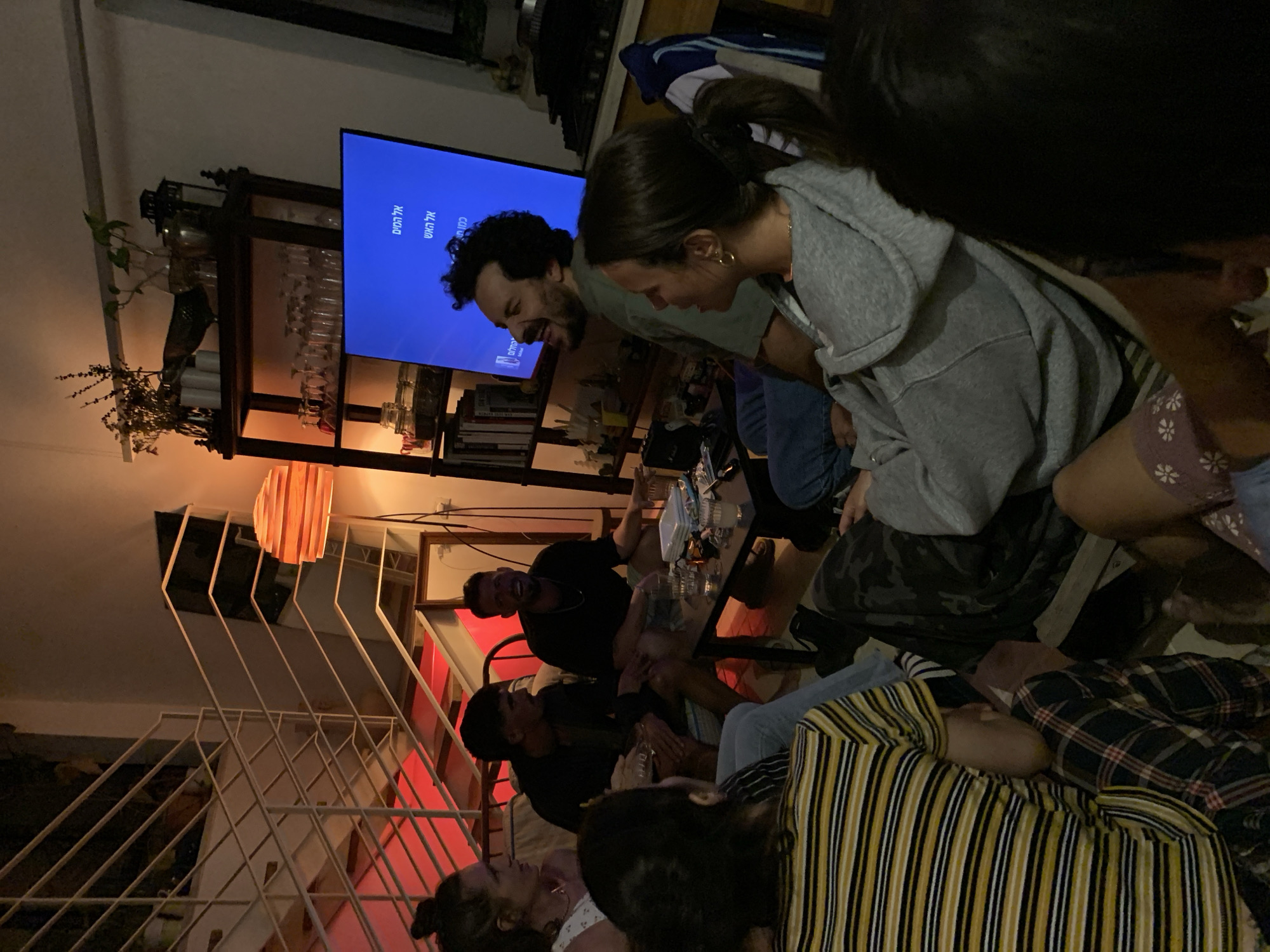 The image captures a lively indoor social gathering, likely in a living room or common area, during the evening or night, indicated by the dim, warm lighting. Several individuals are present, most of whom are seated on the floor around a low table. The atmosphere appears joyful and relaxed, with many subjects seen laughing and interacting.
In the mid-ground, a man with curly dark hair and a beard, wearing a light-colored shirt, is laughing while looking towards a large flat-screen TV. Next to him, a woman with dark hair tied back in a ponytail, wearing a grey hoodie, is also smiling or laughing, looking downwards. Further down, another man in a black shirt is visible, also laughing, seated near the table where drinks and possibly a board game are laid out. Other people are partially visible in various seating positions, some with their backs to the camera, including one person in a yellow and black striped shirt in the foreground.
To the left, a wooden shelving unit or bar cart is positioned below and beside the TV, adorned with various glasses, bottles, and a green plant. Books are also visible on a shelf beneath the TV. A modern, slatted wooden pendant lamp hangs above a white metal staircase railing, visible in the bottom left. The floor appears to be wooden.
The TV screen displays Hebrew text, which includes "אל האש" (To the fire), "אל המים" (To the water), and "כוח" (Power/Strength), with "אלירם" (Eliram) at the bottom. This text, combined with the given location context, suggests the setting is in Tel-Aviv, Israel. The group seems to be engaged in conversation or an activity together, contributing to a vibrant social scene.