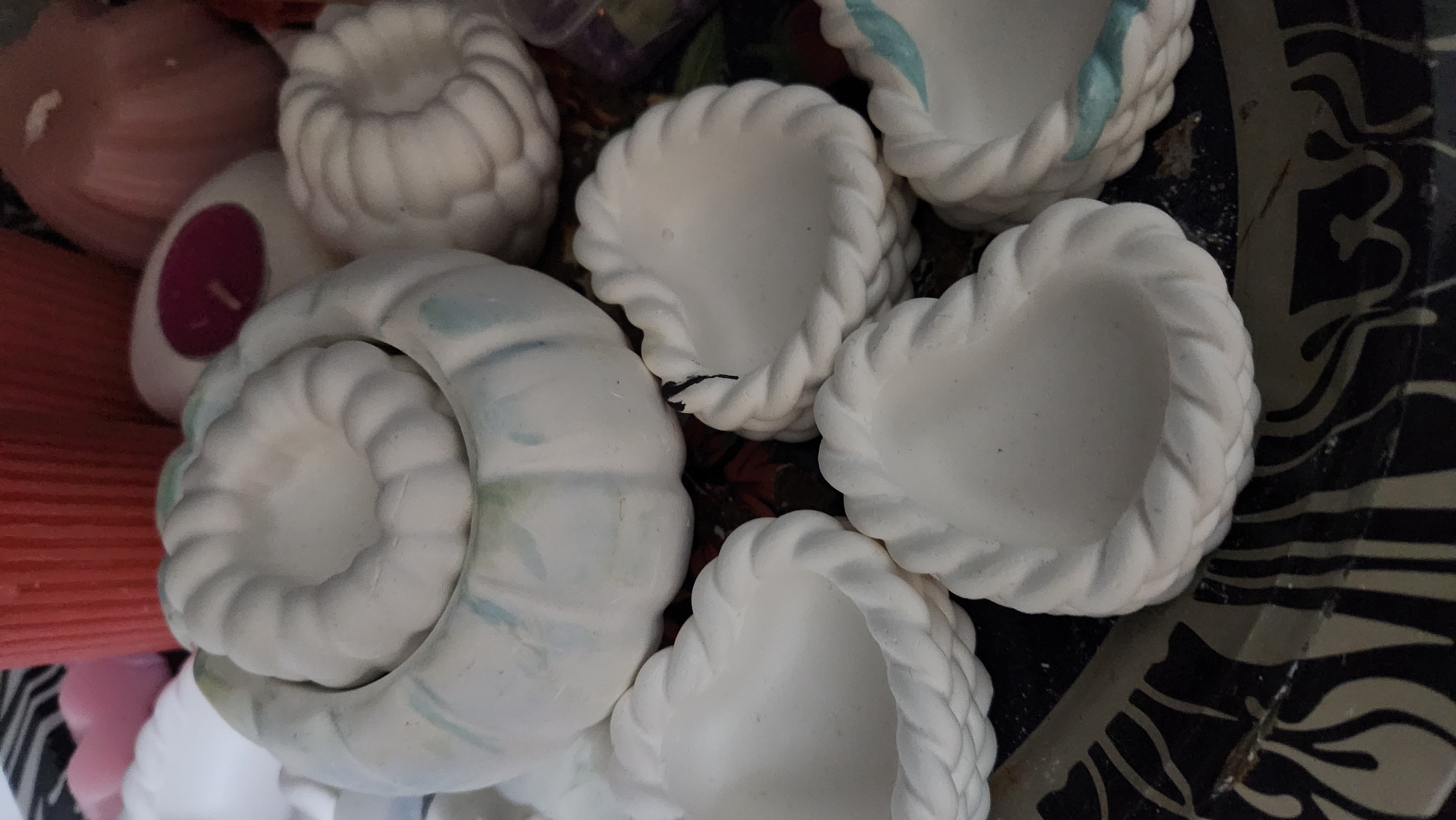 The image shows a collection of white, decorative items that appear to be ceramic or plaster. In the foreground, several heart-shaped dishes with a braided rim are visible. Behind them, a larger, round, pumpkin-shaped dish is present. Another smaller, similar pumpkin-shaped dish is positioned to the right. In the background, there are some red and pink candles, one with a dark pink insert. The items are placed on a dark tray with a decorative pattern. The scene is an indoor setting. There is no information in the image to suggest a location in Buynaksk, Russia.