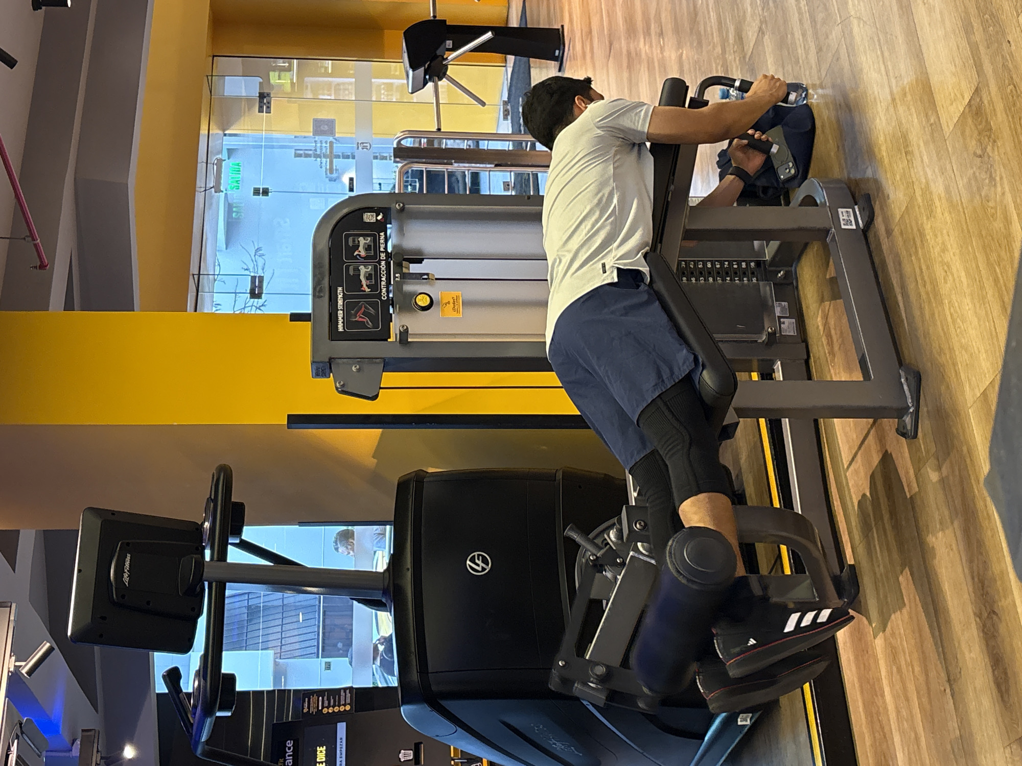 The image shows a man performing a leg curl exercise on a weight machine in a gym. He is lying face down on the padded bench, with his legs bent at the knees and the padded roller resting against his ankles. His hands are gripping the handles of the machine. The gym appears to be well-equipped with other exercise machines and has a modern aesthetic, with yellow walls and wooden flooring. A glass door with the word "SALIDA" (exit) visible in green lettering is in the background, suggesting the location is San Isidro, Peru. There is also a turnstile visible near the exit. The man is wearing a light-colored t-shirt, dark shorts, black compression leggings, and black and white athletic shoes. A blue gym bag and a water bottle are on the floor next to the machine.