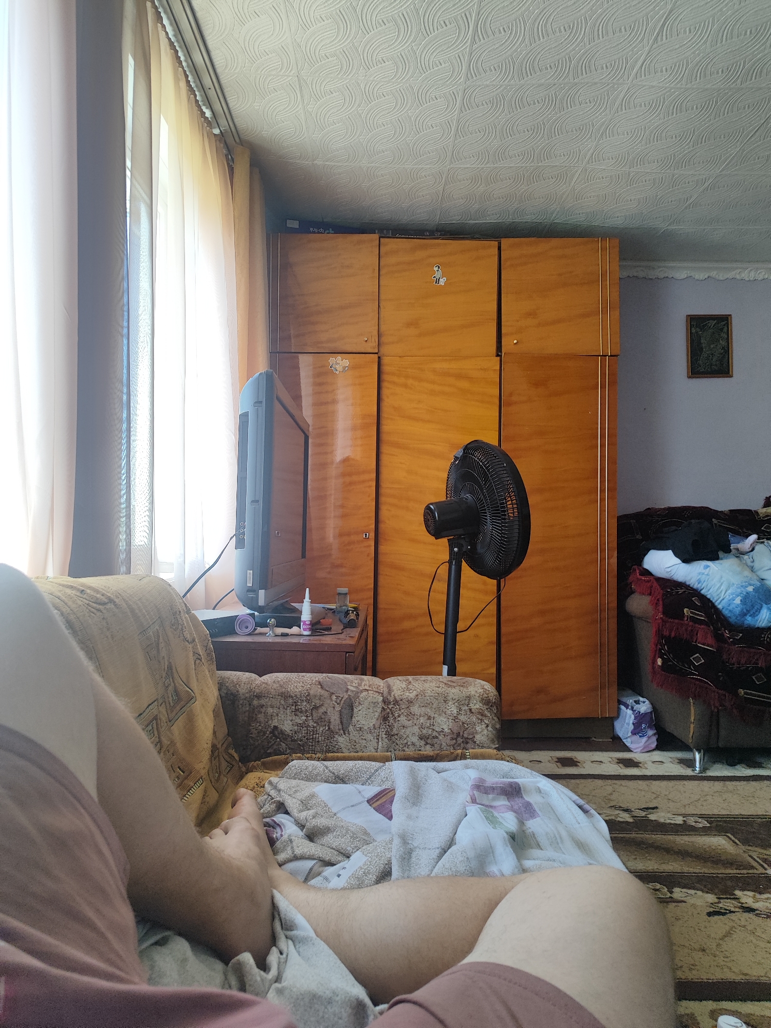The image depicts a domestic indoor scene, likely a living room, captured from a low-angle perspective. The focus is on a person's legs and feet, resting on a couch covered with blankets. The environment suggests a modest living space. To the left, sheer curtains cover a window, allowing natural light to filter into the room. In the background, a large, wooden wardrobe stands prominently. A vintage television set is positioned on a small cabinet to the left of the wardrobe, and a black standing fan is visible in front of the wardrobe. The floor is covered with a patterned rug. A small framed picture hangs on the wall behind the fan. The overall impression is that of a lived-in, possibly older, home. There is no specific indicator within the image that places it in Ridder, Kazakhstan, or any other specific location.