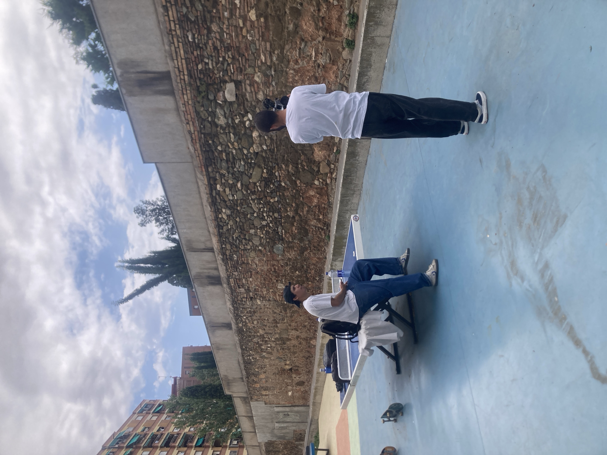 In Sant Feliu de Llobregat, Spain, two men are present on a blue-surfaced outdoor recreational area. One man, seated on a ping pong table, wears a white t-shirt, blue jeans, and a black baseball cap. He is holding a phone in his right hand and has a backpack and water bottle on the table beside him. The other man, standing and facing away from the camera, wears a white t-shirt and dark trousers, holding a video camera. The background features a weathered brick wall, a concrete retaining wall, and residential buildings. The ground is wet in patches, and two skateboards are visible on the blue surface near the ping pong table.