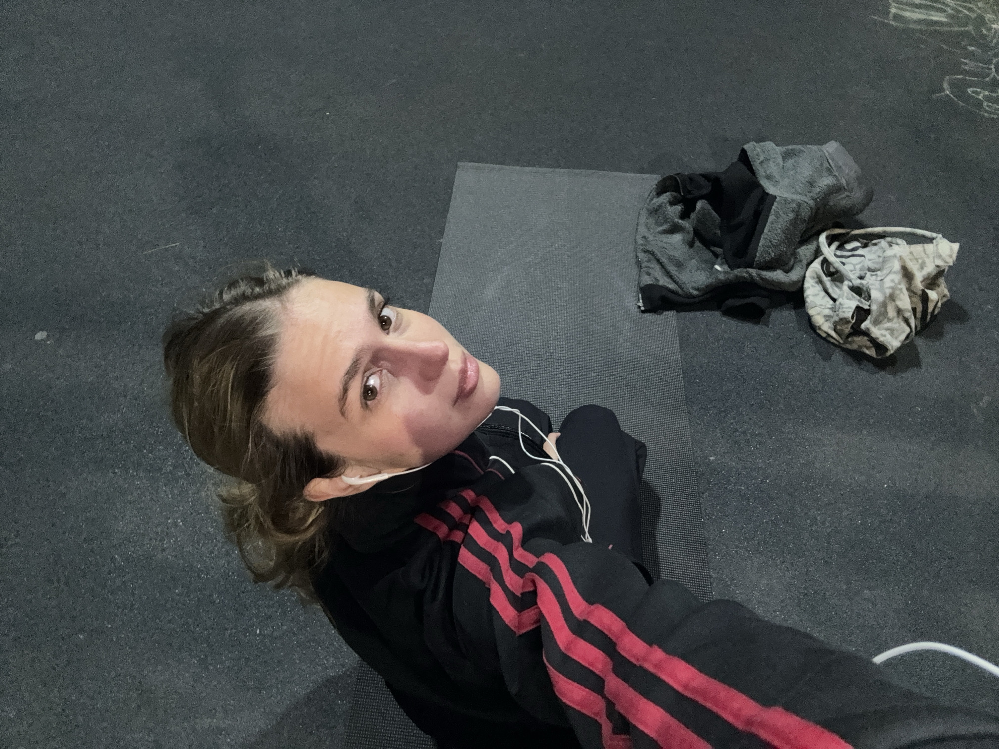 A young woman is captured in a high-angle shot, looking up towards the camera. She has light brown hair, pulled back, and is wearing a black athletic jacket with prominent red stripes on the sleeves. White wired earphones are visible in her ears and extending down. Her expression is neutral, perhaps contemplative or tired.
She is situated on a dark grey exercise mat, which is placed on a textured dark grey floor, likely rubber or carpet, characteristic of a gym or fitness studio. To her right, off the mat, lies a grey hoodie or jacket and a crumpled piece of white or light-patterned clothing, possibly a t-shirt or towel. The overall lighting is somewhat dim, suggesting an indoor setting, possibly during the evening or night. The woman appears to be resting or taking a break from a workout session. The general setting suggests an athletic or fitness activity in Tbilisi, Georgia. There is no visible text in the image.