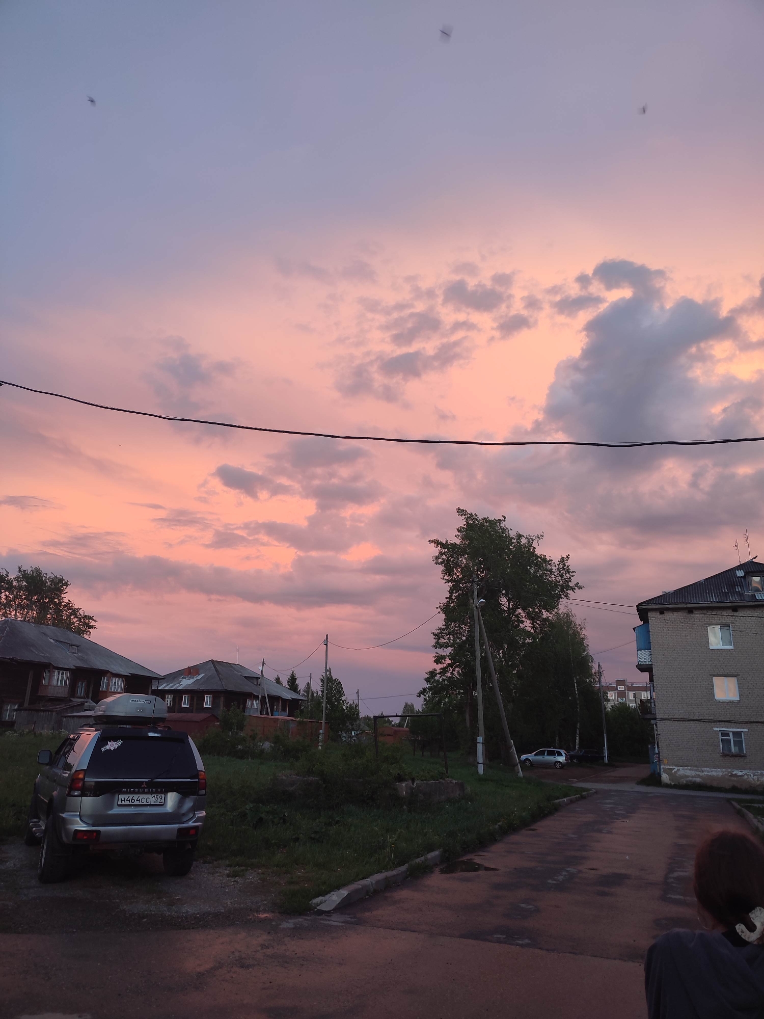 The image shows a street scene in Gremyachinsk, Russia, at dusk. The sky is a mix of pink and purple hues with scattered clouds. Overhead, a power line stretches across the sky. A silver Mitsubishi SUV with a roof rack is parked on the left side of the frame. The license plate is H464CC 159. Behind the SUV are wooden houses with dark roofs. To the right of the SUV is a grassy area with vegetation. A paved street leads into the distance, where a white car is parked. On the right side of the street is a brick apartment building with a dark roof. A person with dark hair tied up is partially visible in the lower right corner, facing away from the camera.