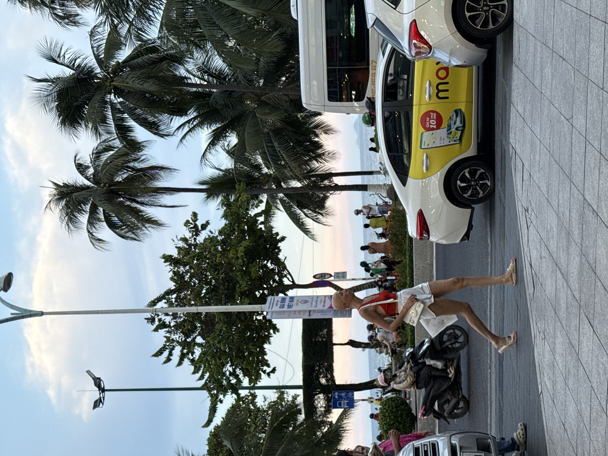A medium shot captures a street scene in Nha Trang Ward, Vietnam, during what appears to be twilight or overcast conditions. The foreground shows a paved sidewalk with a grey, grid-patterned surface. A woman with a shaved head, wearing a red bikini top and white shorts, is walking across a street towards the right of the frame. She carries a white handbag on her left shoulder. Her right leg is extended forward, mid-stride, and she wears light-colored sandals.
To the left, a silver car is partially visible, with a person in blue jeans and beige Crocs standing next to it. Further left, a motorcycle is seen on the road, ridden by a person wearing a beige jacket and a pink cap. Behind the woman, the street is populated with other people, some standing and some on motorcycles.
The background reveals palm trees and other lush green foliage. A tall street lamp with a curved arm and a light fixture stands prominently on the left side of the frame, with another similar lamp further in the distance. A white van is parked on the right side of the street, and to its right, the rear of a white car is visible. A yellow taxi, identified by the word "max" and an advertisement featuring a car and text in Vietnamese, is positioned between the van and the white car. The sea and a distant hazy landmass can be seen beyond the street and trees. A banner with Vietnamese text is attached to a pole near the street lamp on the left. The overall atmosphere suggests a bustling urban environment near a coastal area.