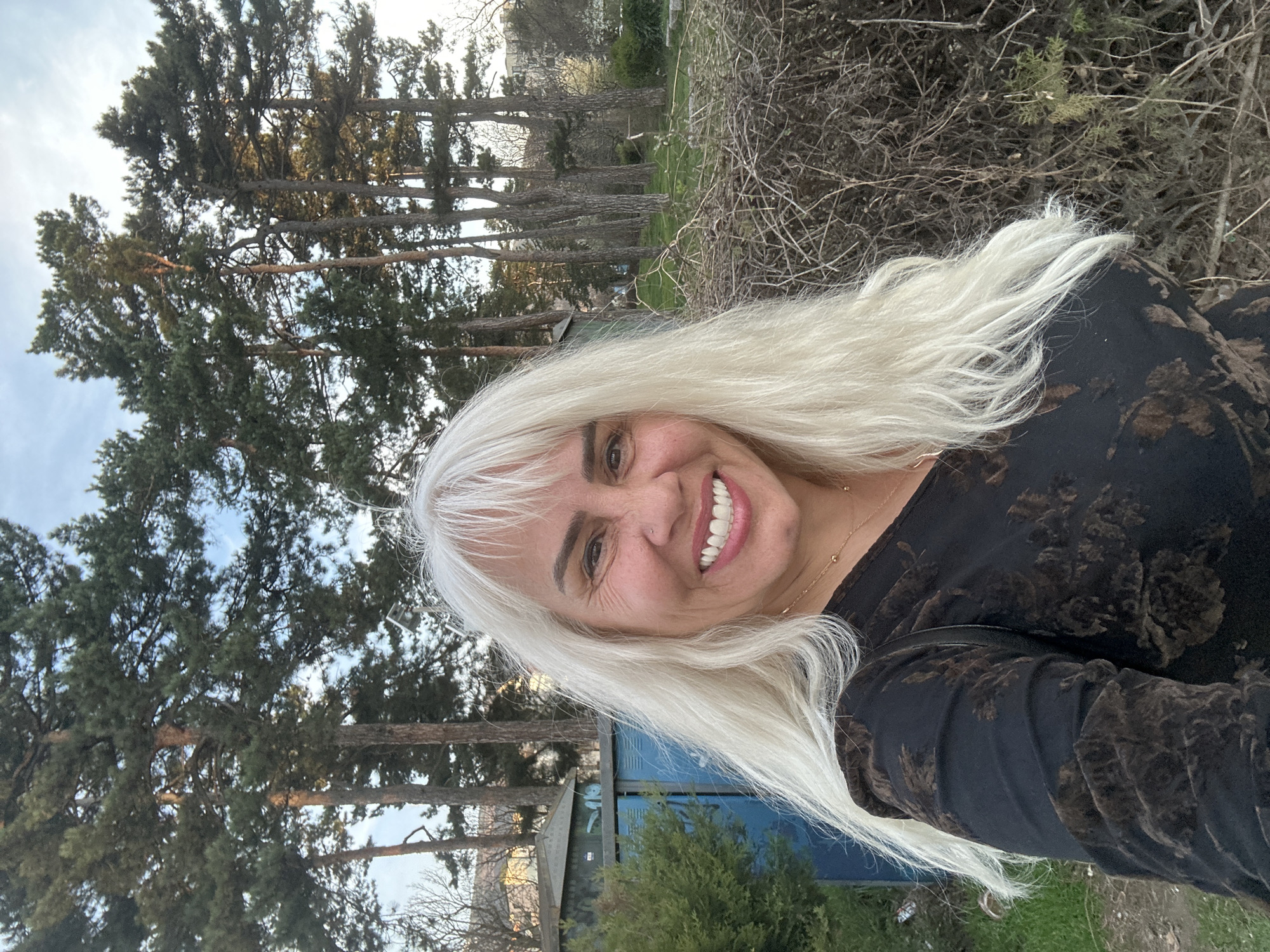 A medium shot captures a smiling woman with long, straight, platinum blonde hair. Her dark floral-patterned shirt suggests the cooler temperatures. The backdrop reveals a landscape including tall, thin trees, a small, blue portable toilet unit and a pile of dry shrubbery. Given the vegetation and the presence of urban amenities like the portable toilet, it is likely to be a park or green space within the city. The image seems to have been taken outdoors, at dusk. Given that the request is to identify if this is in Sofia, Bulgaria, it is difficult to determine definitively if this image was taken in the city of Sofia.