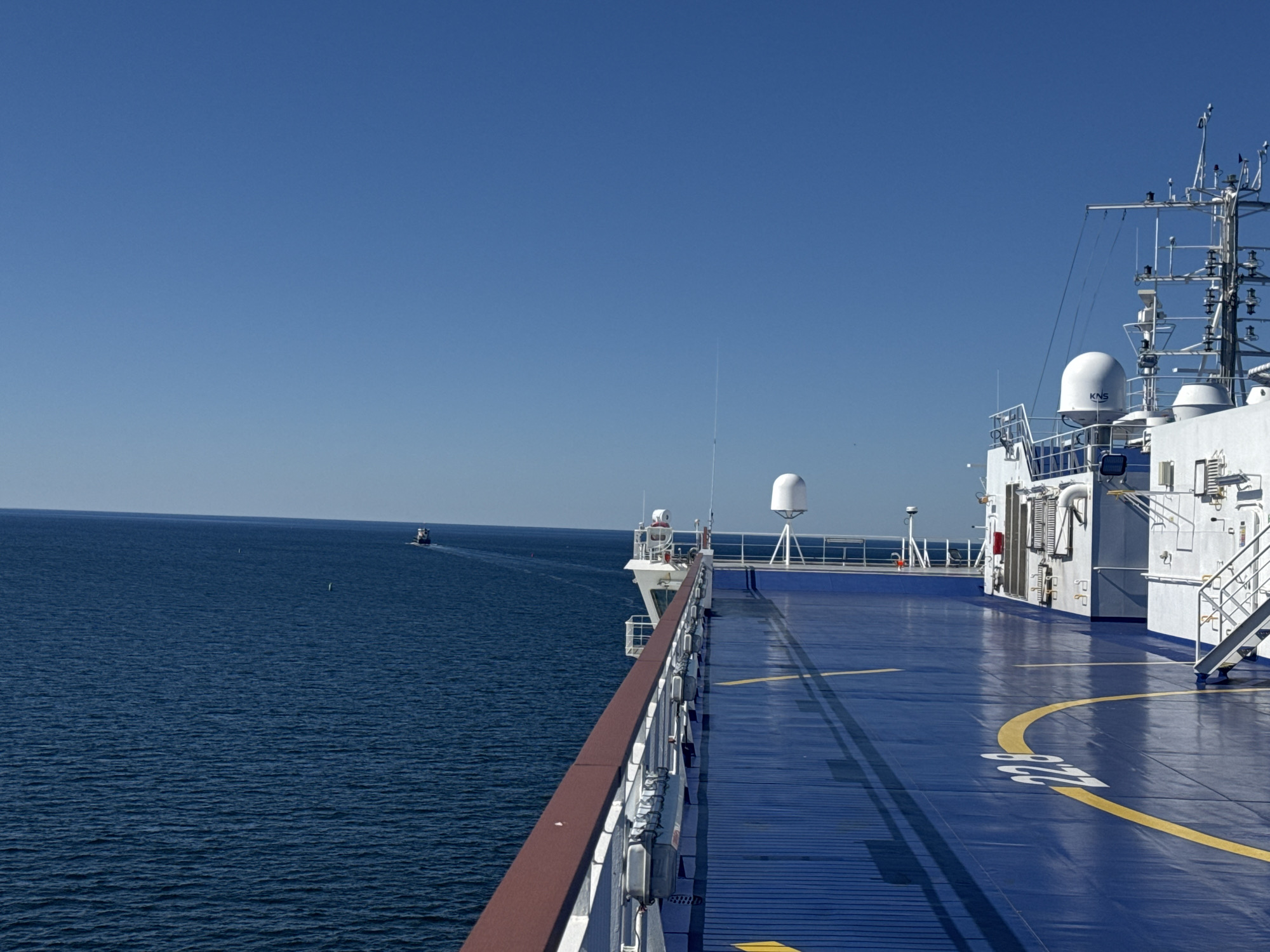 The image shows the deck of a white ship with a blue surface, marked with yellow lines including a circular pattern and the number "20". The ship's railing, with a brown top, runs along the left side of the deck. The ship's superstructure, featuring various antennas, domes (one labeled "KNS"), and equipment, is visible on the right. The scene is set in the open sea under a clear blue sky. In the distance, on the left, a smaller boat is visible on the water. The location is identified as Drottningskär, Sweden.
