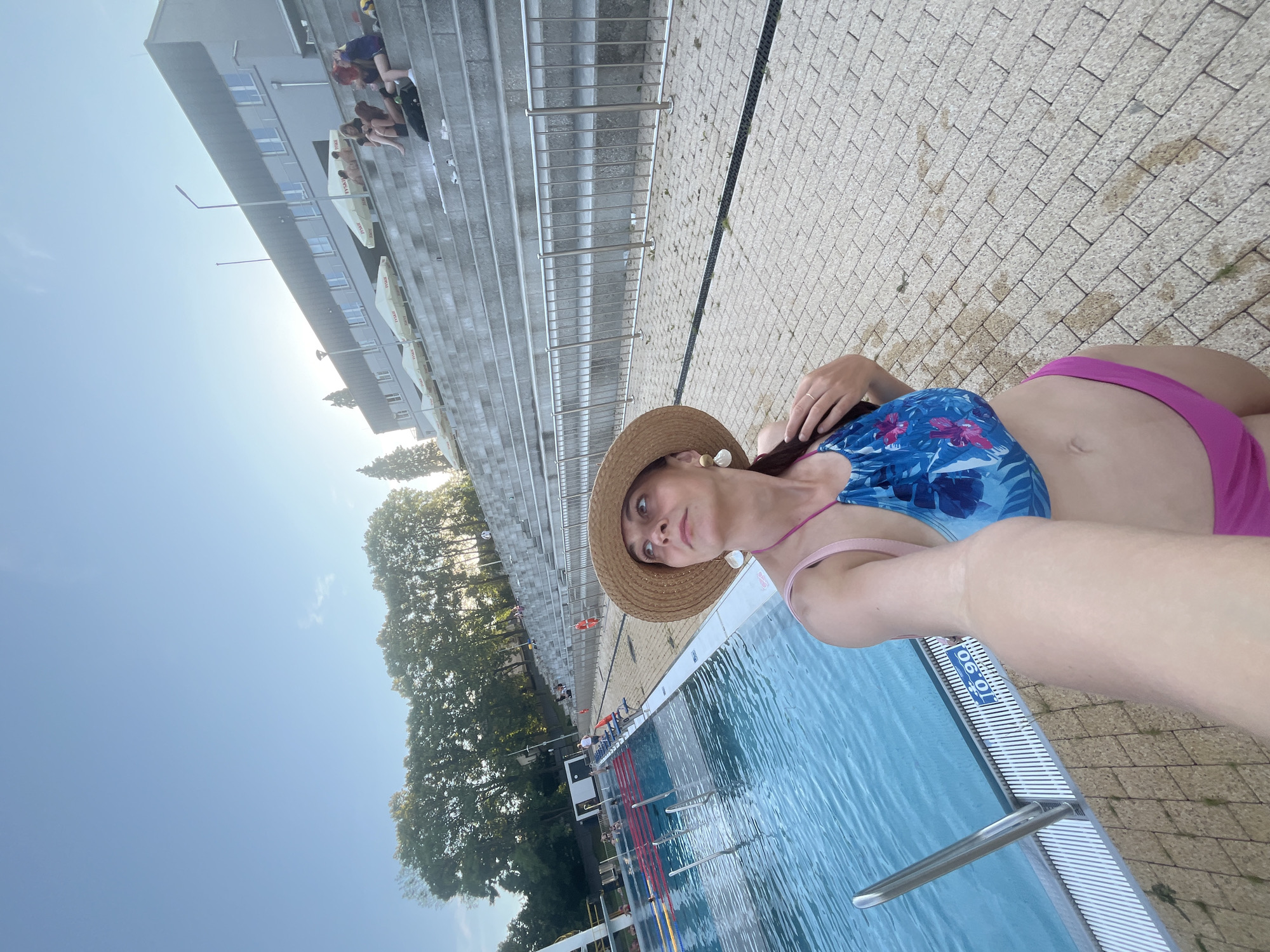 A woman in a blue and pink floral bikini and a straw hat stands near the edge of a swimming pool, looking up and to the right. She is holding her right hand to her hair. To her left is a swimming pool with lanes marked. Behind her are tiered concrete bleachers with people sitting on them. A building is visible at the top of the bleachers. The sky is clear and blue.