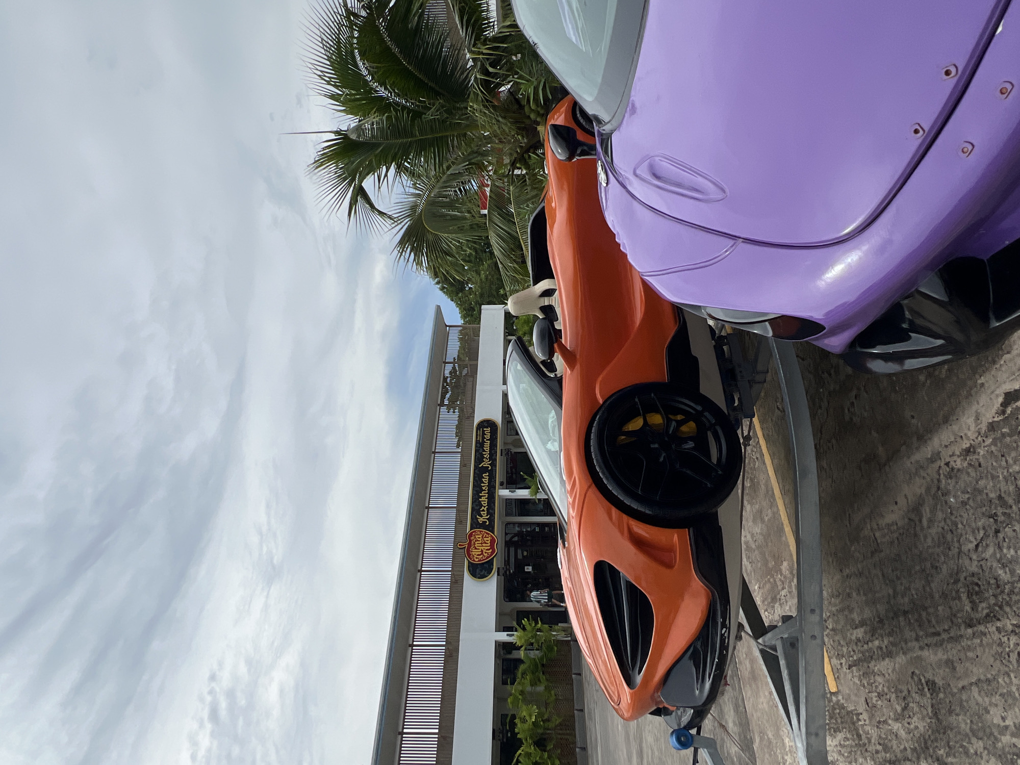 Two McLaren sports cars, one orange and one purple, are parked on a trailer in front of a restaurant called "Alma Kazakhstan Restaurant" in Chalong, Thailand. The orange car is in the foreground and is a convertible, with its top down. The purple car is partially visible behind the orange car. The restaurant has a modern design with a wooden facade and is surrounded by palm trees. The sky is cloudy. There are no people clearly visible in the image.