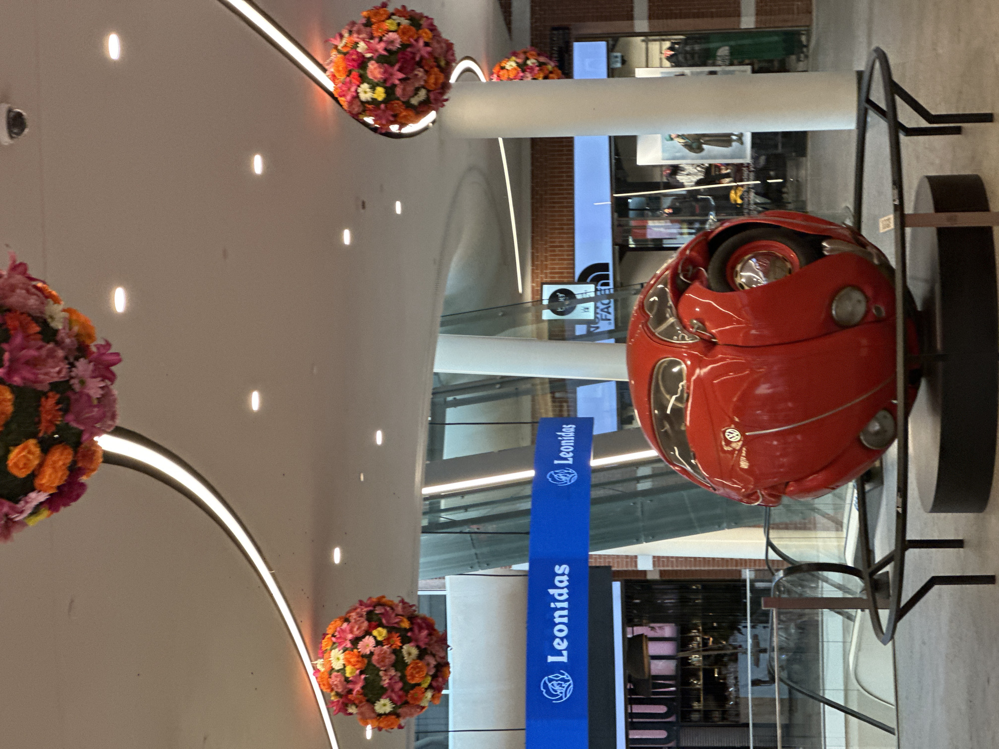 An indoor scene features a red, spherical sculpture resembling a stylized car, possibly a Volkswagen Beetle, positioned on a black circular platform. The sculpture is illuminated by ambient lighting and spotlights on the ceiling. Three large, spherical floral arrangements, comprised of pink, orange, and yellow flowers, are suspended from the ceiling, adding a vibrant splash of color to the upper part of the image. A blue banner with the "Leonidas" logo is visible in the background. The environment appears to be a shopping mall or public indoor space, indicated by the presence of escalators and storefronts in the background. A security camera is mounted on the ceiling. No people are clearly visible in the foreground.