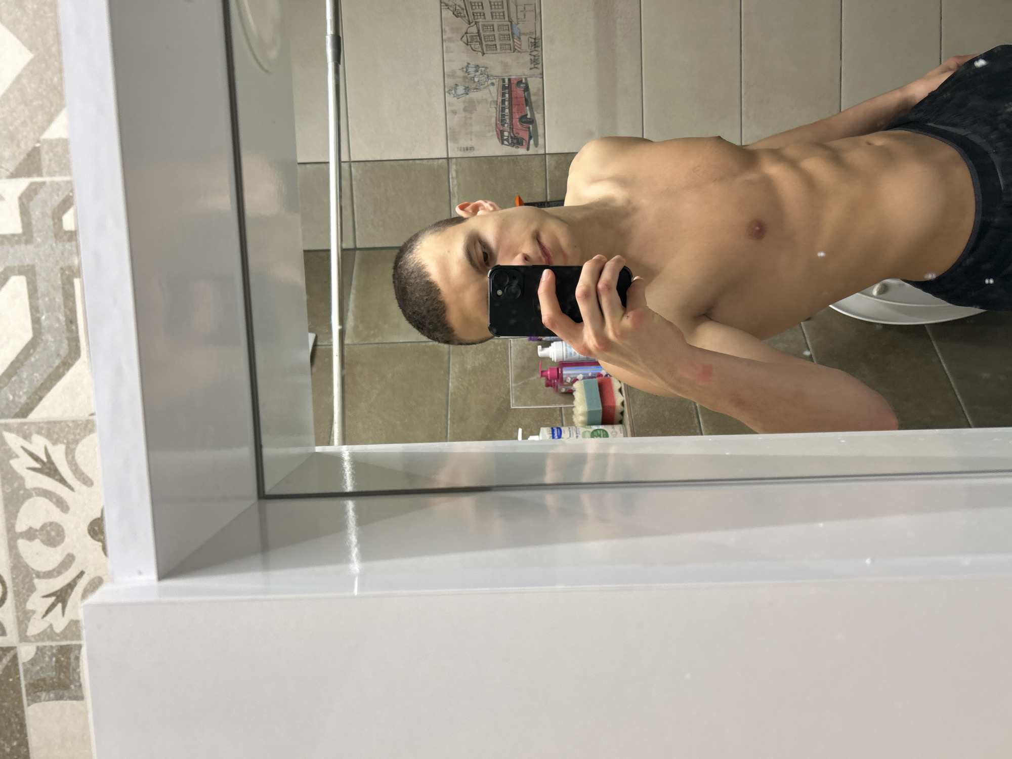 A shirtless man is taking a selfie in a bathroom mirror. The man has short dark hair and is wearing dark colored bottoms. The bathroom has tiled walls, and there is a shelf with toiletries behind the man. The picture on the wall shows an image of a city, with a red double decker bus. The location is unknown, and there is no indication that it is in Burgas, Bulgaria.
