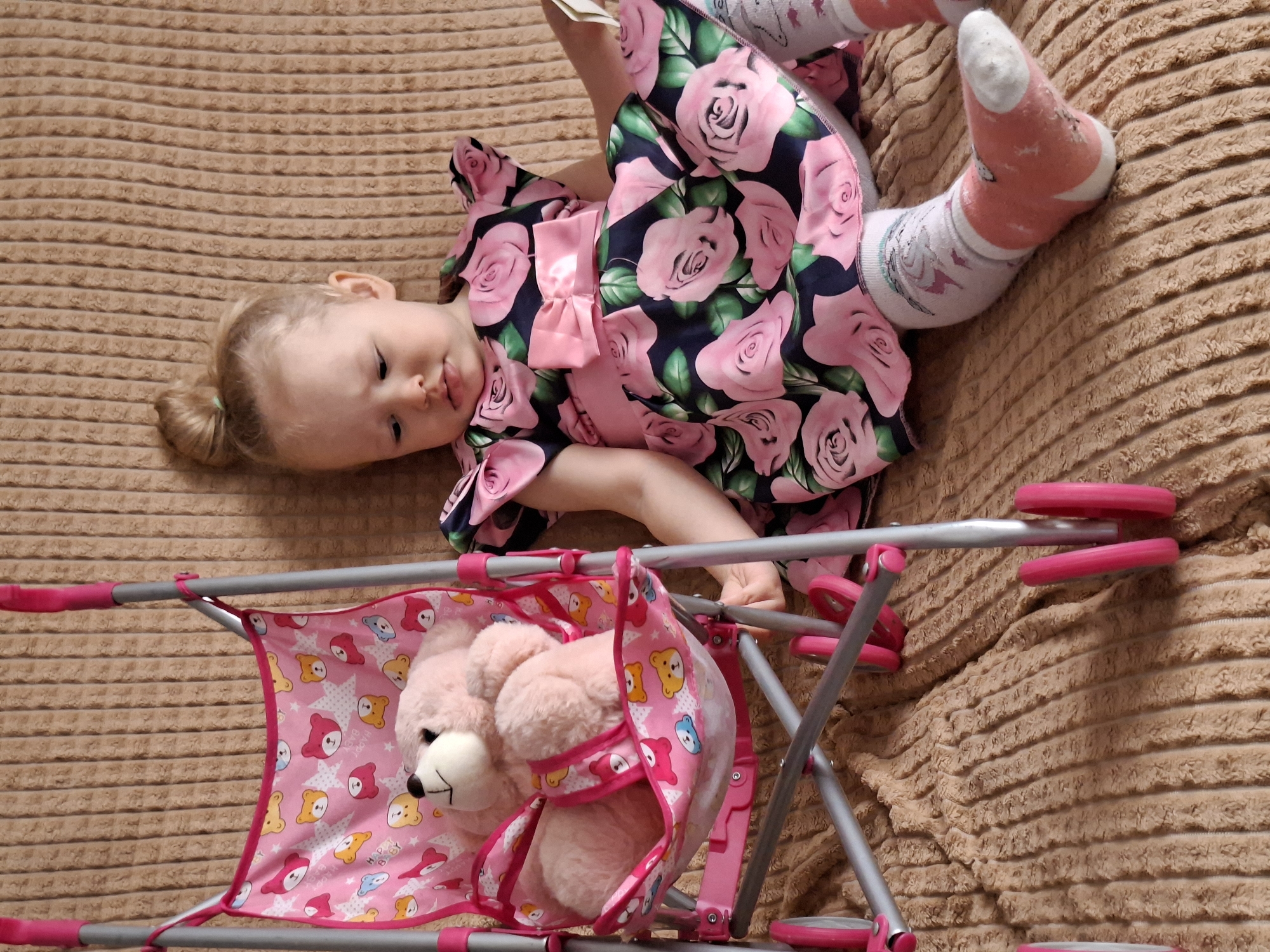 A young child, dressed in a pink floral dress and patterned socks, is lying on a textured brown surface, likely a bed or blanket. The child is near a pink toy stroller with a plush teddy bear inside. The location appears to be indoors.