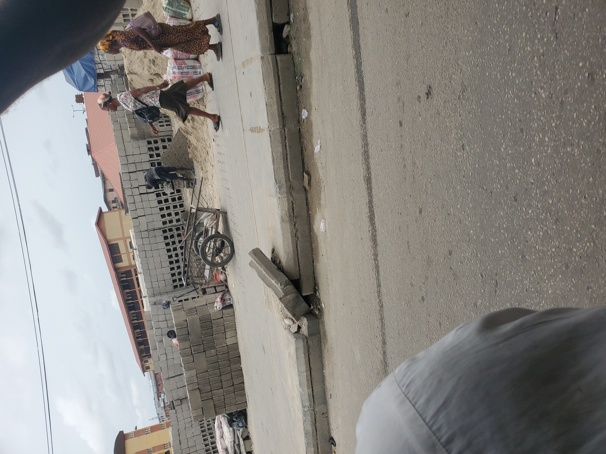 This image captures a bustling street scene in Alaiyabiagba, Nigeria, during the daytime under an overcast sky. The view is partially obscured by what appears to be the photographer's shoulder or clothing in the bottom right corner.
On the right side of the frame, there's a paved road. To its left, a concrete sidewalk runs parallel, with a broken curb separating it from the road. Various small pieces of white debris and trash are scattered on the ground near the curb.
The left side of the scene is dominated by large stacks of concrete blocks, suggesting a construction material depot or an ongoing building project. Piles of sand are visible behind and around these blocks. Further back, buildings with red and yellow/brown roofs and facades are visible. Utility lines cross the sky in the upper left corner.
Several people are present in the scene. Two women are walking on the sidewalk: one wears a leopard-print dress and a yellow headscar, carrying a purple bag, while the other has a patterned top, dark shorts, a light head covering, and a black shoulder bag. Near the stacks of blocks, a person in dark clothing is bent over, seemingly working with the blocks or sand. The frame of a handcart or wheelbarrow is also visible among the blocks.
The atmosphere appears to be one of everyday activity and commerce related to construction. No specific text is clearly legible on any objects within the image.