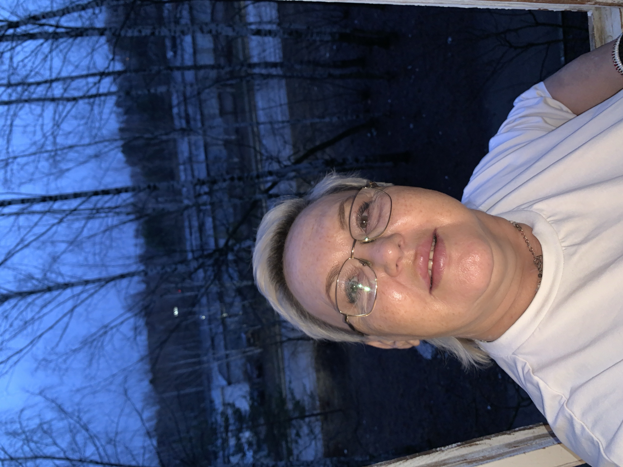 A woman with short, platinum blonde hair and glasses is standing near a window. The background shows a blurred scene of trees, likely outdoors. The photo was taken in the city of Chaikovsky, Russia.