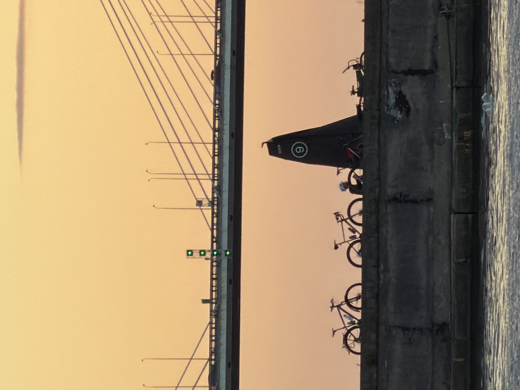 The image depicts a scene at sunset in Saint Petersburg, Russia. On the left, a section of a cable-stayed bridge is visible, with its numerous cables extending upwards against a hazy, orange sky. A traffic light with three green signals is also present on the bridge.
The right side of the image shows a concrete embankment, with water reflecting the sky on the far right. Along the embankment, a dark, triangular tarp or sail with a white circle containing the number "6" is propped up. Several bicycles are lined up behind the tarp, some leaning against it and others standing on their own. One cyclist, wearing a white helmet, is visible near the bottom of the frame, tending to a bicycle. The overall mood is calm, with the soft light of sunset casting long shadows and creating a serene atmosphere. The setting suggests a waterfront area, possibly a promenade or a park near a bridge and a body of water.