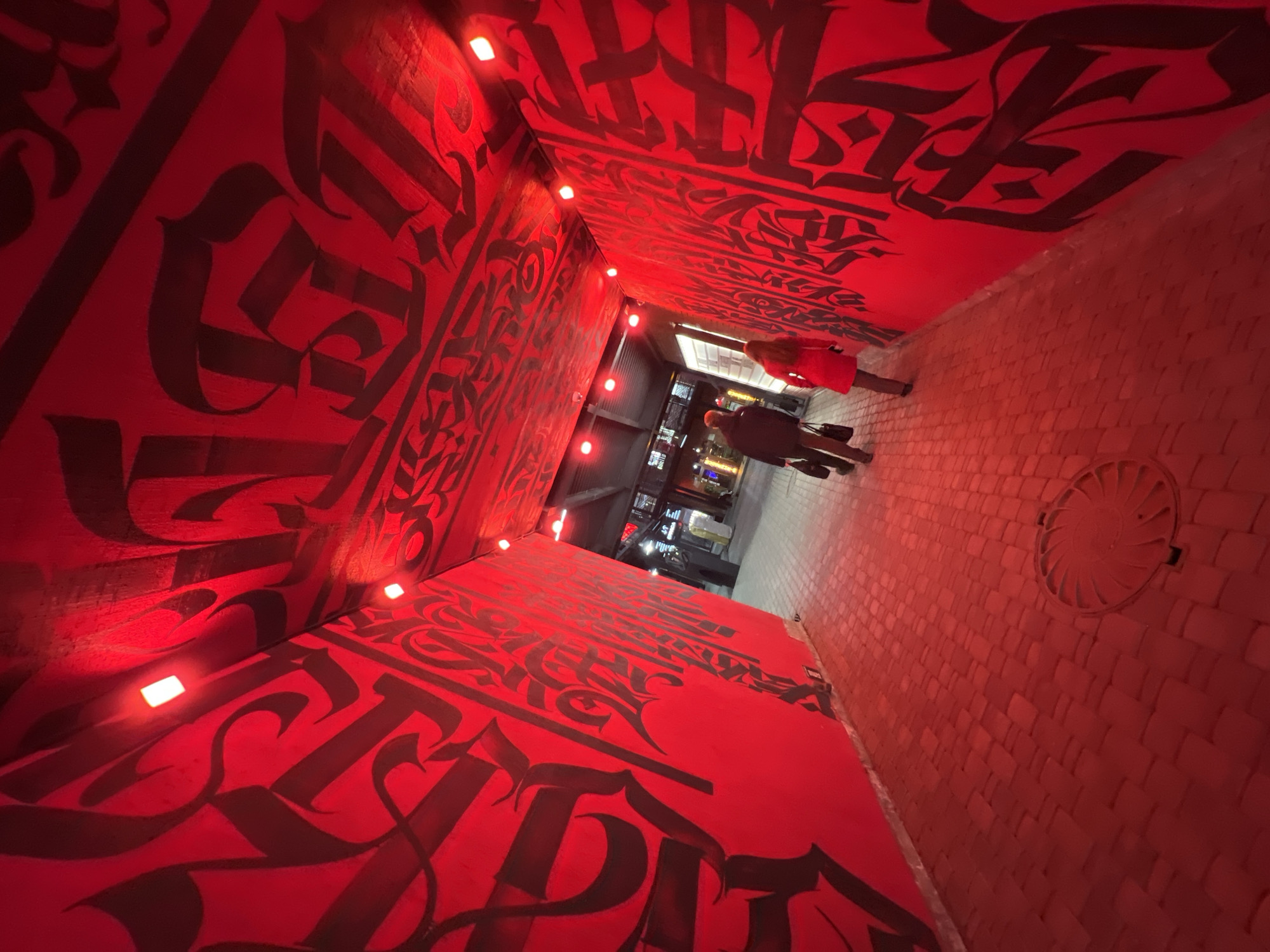 The image shows an alley in Saint Petersburg, Russia. The alley walls and ceiling are painted red with black gothic script. Small, square LED lights line the top of the walls. The alleyway is paved with red brick. Two people are walking towards the end of the alley, a person wearing a red coat and a person wearing a dark coat. A sewer grate is visible on the right side of the brick walkway. Outside the alley entrance, there is a cityscape view, with buildings and lights visible.