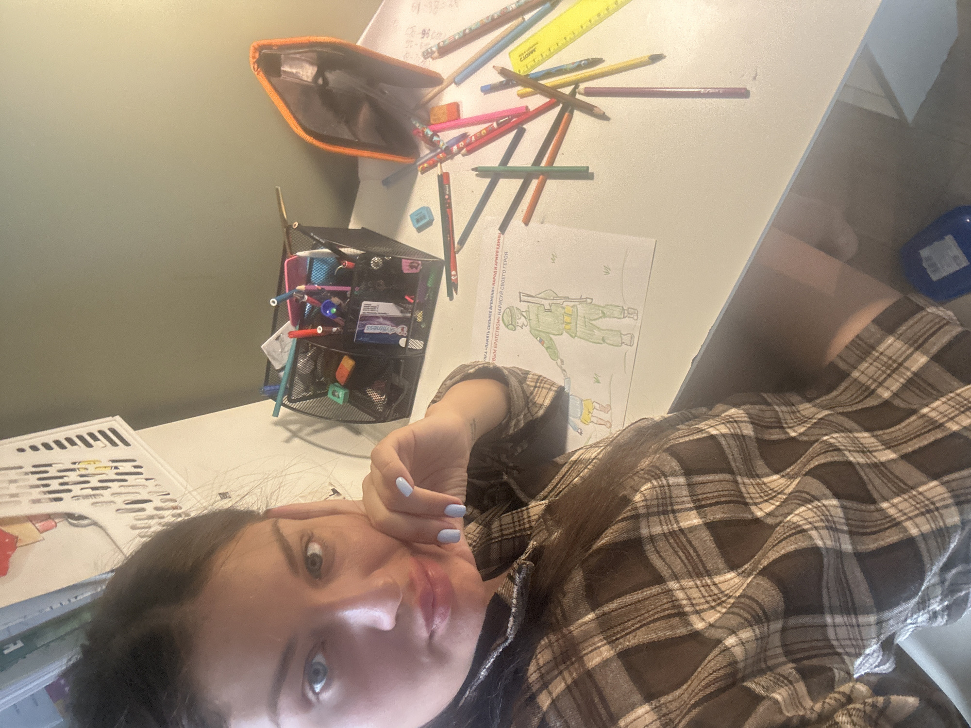 A selfie taken by a young woman in a plaid shirt, possibly at home, shows part of her face and upper body along with a cluttered desk. The desk holds a pencil case, numerous colored pencils and pens, a ruler, a container of writing implements, and papers. A drawing of a soldier can be seen on one of the papers. A text, partly readable as "ПАМЯТЬ СИЛЬНЕЕ ВРЕМЕНИ - НАРОД И АРМИЯ ЕДИНЫ - НАРИСУЙ СВОЕГО ГЕРОЯ" is also on the drawing. "skyfitness" is visible on one of the items. The OCR hints indicate that the text on the image is Russian. There is nothing in the image that locates it to the city of Khimki, Russia.