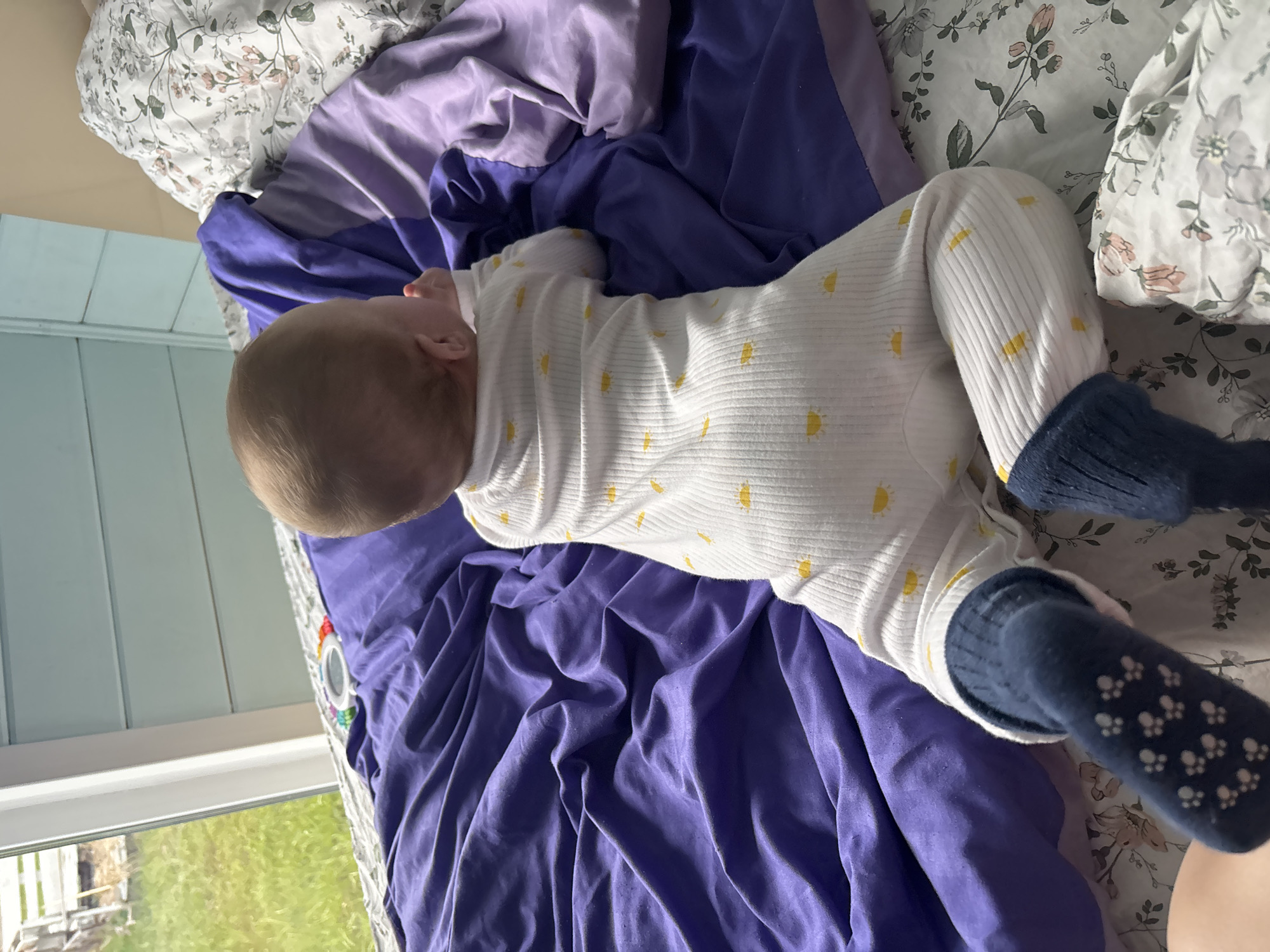 A baby in a white onesie with yellow suns and blue socks lies on a bed with a purple blanket and floral sheets. The location cannot be determined from the image.