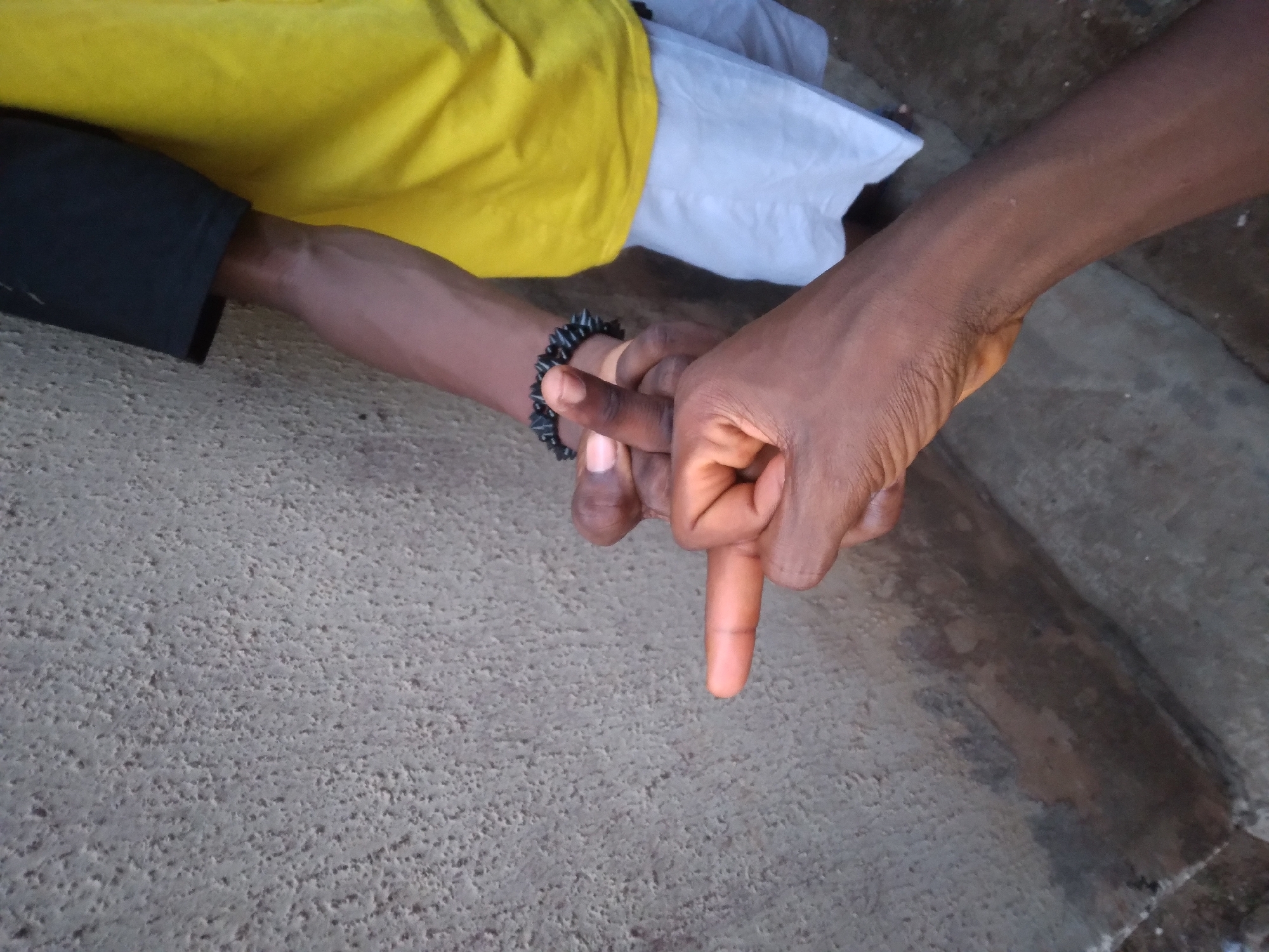 Two dark-skinned hands are intertwined. One hand, adorned with a spiked black bracelet, has its index finger pointed down, touching the index finger of the other hand, forming an "L" shape. The background consists of a concrete surface, a yellow shirt, a black sleeve, and a white cloth. The location of the image is Onitsha, Nigeria.