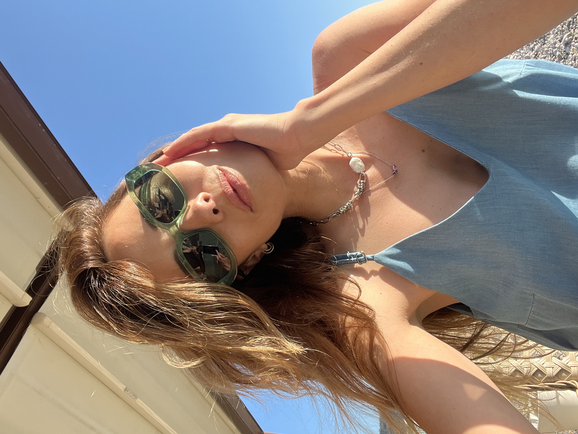 A female subject is captured in a close-up selfie, posing outdoors under bright sunlight. Her head is tilted slightly, and her left hand rests against her cheek and temple. She wears large, light green-framed sunglasses that reflect a mobile phone screen and hands, indicating she is taking the photograph. Her long, reddish-brown hair, styled with loose waves, falls over her shoulders and is illuminated by the sun, showing various highlights and some flyaways.
She is wearing multiple silver-toned necklaces; one features a prominent, irregular white pearl, and another, thinner chain holds a small, possibly purple, gemstone pendant. A small hoop earring is visible in her left earlobe. Her attire consists of a light blue, denim-look camisole or dress with thin straps and a V-neckline.
The background features a clear, deep blue sky on the right side. To the upper left, part of a white and brown retractable awning or canopy structure is visible, suggesting a shaded seating area. The lower right portion of the image shows a ground covered in small, light-colored pebbles, characteristic of a pebble beach. The strong, direct sunlight casts defined shadows and highlights on the subject's face, arm, and collarbone. The overall scene is consistent with a coastal setting in the city of Bar, Montenegro.