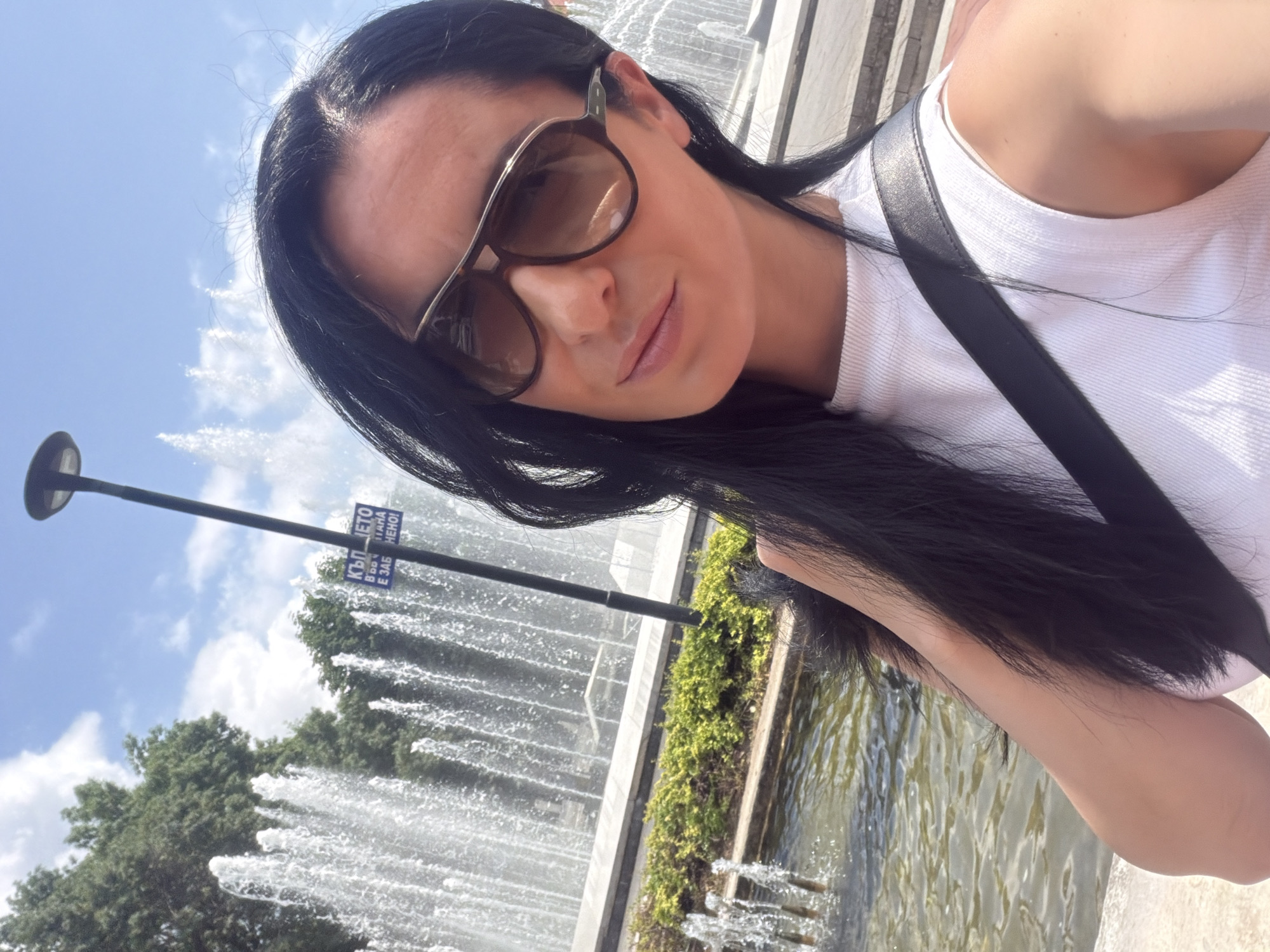 This image is a bright, sunlit selfie of a young woman taken outdoors in Pleven, Bulgaria. The woman has long, dark hair and is wearing stylish dark sunglasses and a white ribbed sleeveless top with a black strap (likely a bag strap) crossing her shoulder. Her expression is calm and neutral, with her lips slightly parted.
The background prominently features a large fountain with multiple jets of water spraying upwards, creating a lively aquatic display. Beyond the fountain, lush green trees stand against a clear blue sky dotted with a few white clouds. A black pole, possibly a lamppost, is visible, and attached to it is a blue sign with white Cyrillic text. The legible parts of the text include "КЪЛВЕТО" and fragmented words such as "В ДО", "ТАНА", "Е ЗАБ", and "НЕНО", which are consistent with the Bulgarian language. The fountain is surrounded by concrete or stone structures and green shrubbery, and the shimmering water of its basin is also visible. The bright lighting indicates it is daytime, likely a sunny mid-day or afternoon. The overall scene suggests a leisurely moment in a public park or square setting.