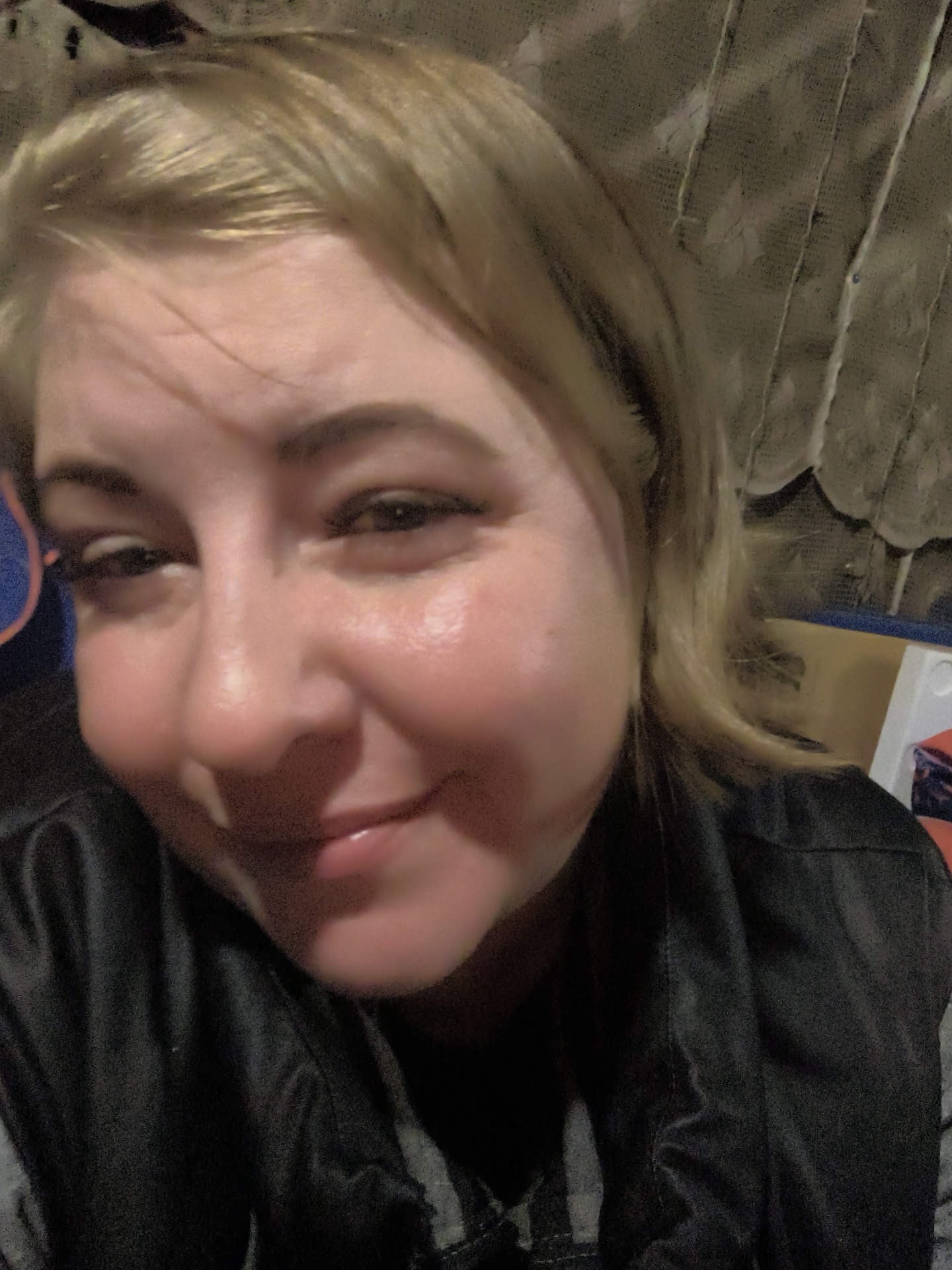 A close-up, low-angle, slightly blurry selfie shows a fair-skinned individual with blonde hair and dark eyebrows. They are wearing a black jacket over a grey garment. The background appears to be an indoor setting with a textured, possibly fabric wall and some white and brown objects on the right. The lighting is dim and uneven, creating highlights on the person's face, particularly on their cheekbones and forehead. The overall impression is that of a casual, informal photograph taken in a dimly lit room. The specific location in the city of Правдист-1 Снт, Russia, is not discernible from the image itself.