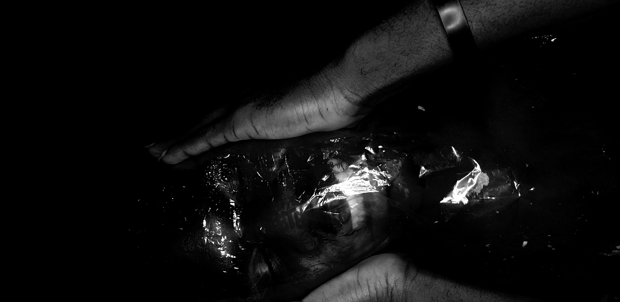 The image is a high-contrast, black and white photograph dominated by darkness and stark illumination. It features two human hands interacting with an object wrapped in crinkled, reflective plastic.
One hand enters the frame from the upper-left, extending downwards with fingers splayed, while the other hand reaches upwards from the bottom-right. Both hands appear to be pressing against or cradling the plastic-covered object, which occupies the central-lower portion of the frame. The upper hand displays visible arm hair and a metallic band or bracelet on its wrist.
Underneath the shiny, crinkled plastic, a dark, indistinct form is visible. This form vaguely resembles a human head or face, though its features are heavily obscured and distorted by the plastic's texture and numerous bright reflections. The background is entirely black, suggesting an absence of light or a very shallow depth of field against a dark surface.
The activity appears to be the handling or examination of the wrapped item. The lack of clear context and the obscured nature of the object create a mysterious and somewhat somber mood. Faint, illegible text and numbers, including "No. 3," can be discerned on the surface of the plastic, indicating it is likely some form of packaging. No specific location cues related to Narayi, Nigeria, are visible within the image.