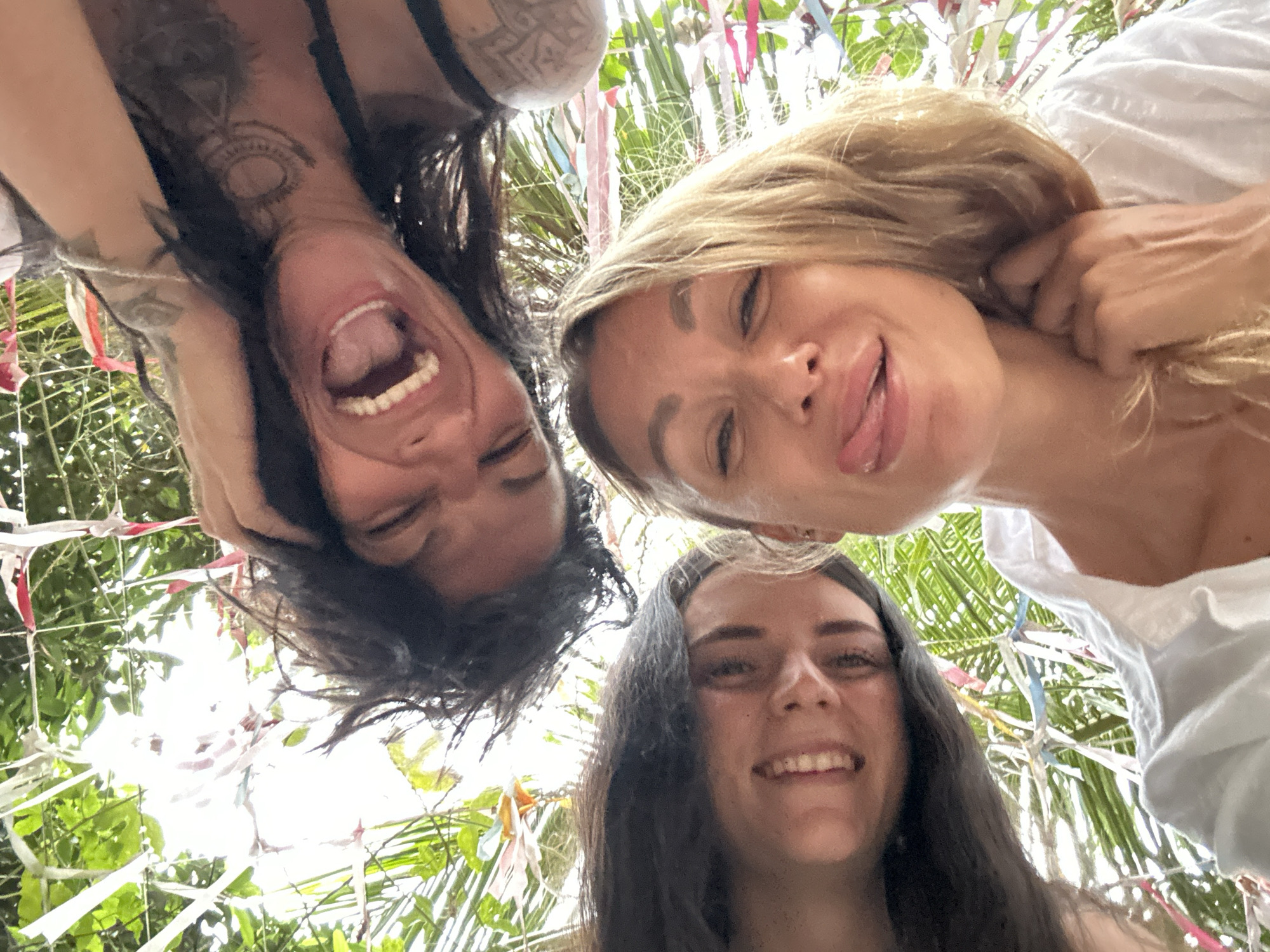 Three young women are clustered together looking down at the camera, likely taken from below. The woman on the left has dark hair, is facing down and slightly to the right, and has her mouth wide open in a laugh; she has visible tattoos on her shoulder and arm. The woman on the right has blond hair, is facing forward and to the left, and has her tongue slightly out in a playful manner; she is wearing a white top. The woman at the bottom has dark hair, is facing forward, and is smiling. Behind them are green plants, and what appears to be a decorative structure with ribbons. Given the prompt, this is likely located in Mũi Né, Vietnam.