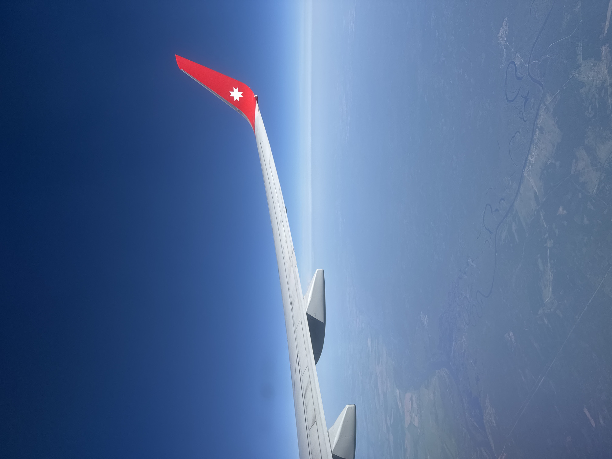 An eye-level outdoor shot from an airplane shows the ground in Russia, with its rural geography, and part of the airplane's wing, during daylight hours.
The left side of the shot is dark blue sky, while the right side shows hazy-blue land in Russia. A river can be seen meandering through the landscape. The left wing of the aircraft is shown against the sky. The wing is predominantly white, with aerodynamic components visible. The tail of the wing has a red section with a white star emblem.
Based on the image's composition and geographic features, it is not possible to specifically identify the exact location as Lyamtsino, Russia. More specific visual or contextual cues related to Lyamtsino are needed to verify the location.
