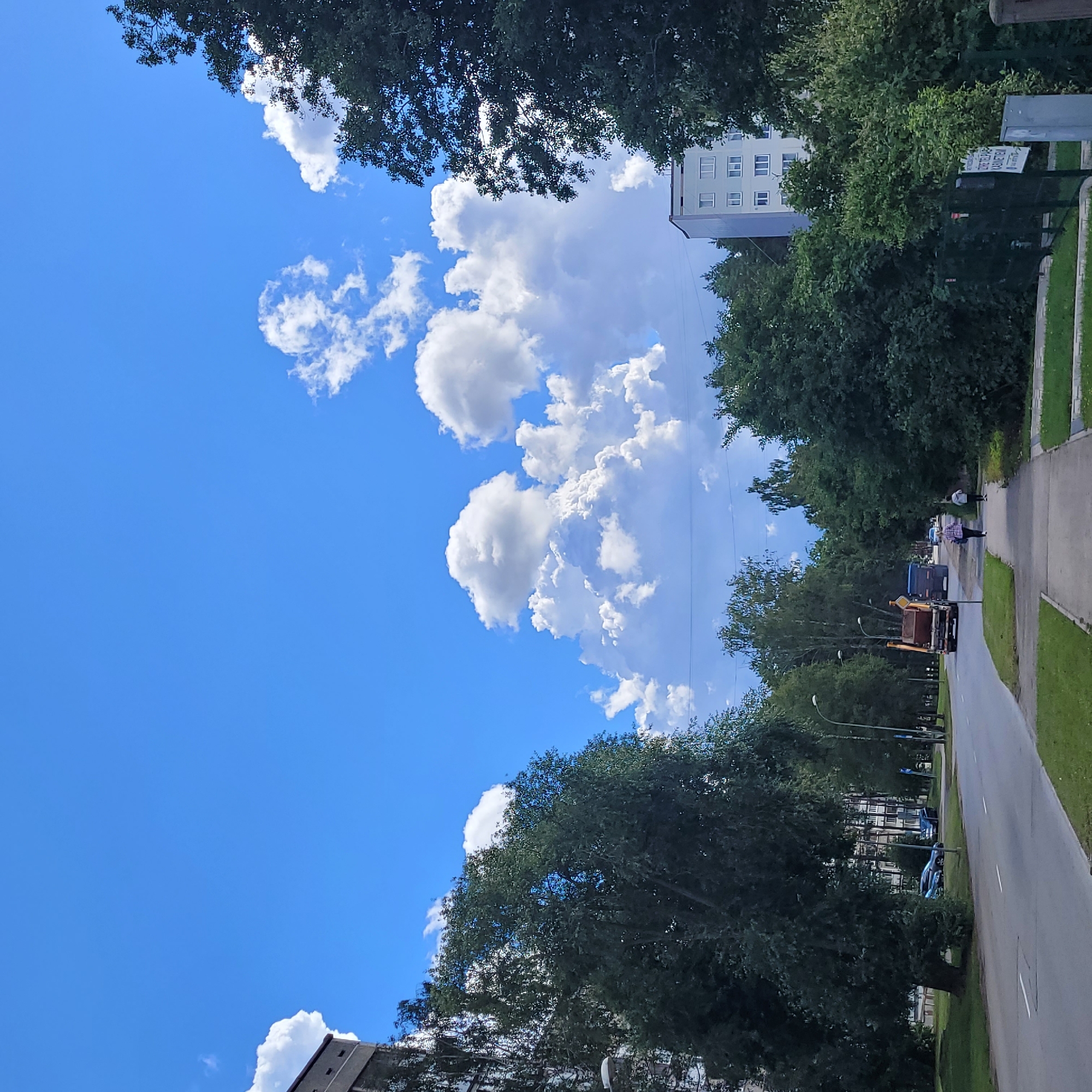 The image depicts a sunny day with a bright blue sky dotted with fluffy white cumulus clouds. In the foreground, a paved road runs horizontally across the scene, bordered by a sidewalk and grassy areas. Several cars are parked along the side of the road. A garbage truck is visible on the road, moving away from the viewer. Two people are walking on the sidewalk.
Behind the road and sidewalk, a dense line of green trees stretches across the middle ground, with some taller trees reaching towards the sky. In the background, apartment buildings are visible, peeking out from behind the trees. The overall impression is of a residential area in a city, likely Jūrmala, Latvia, during a pleasant summer day.