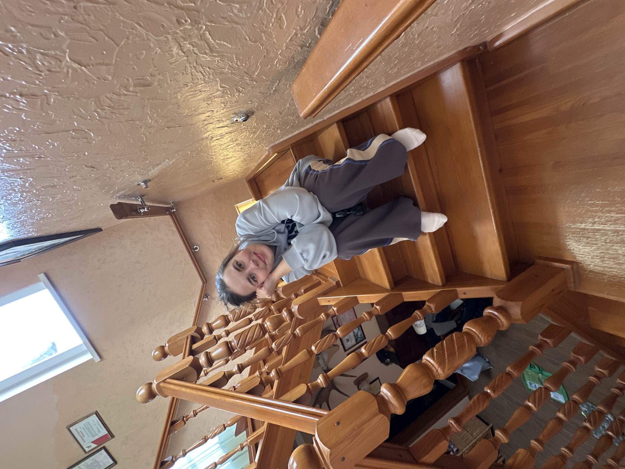 A young woman lies on a wooden staircase inside a building, possibly a residence, in Ilyinsky, Russia. She is wearing a gray sweatshirt and gray sweatpants with a light blue stripe, and white socks. Her head rests on her hand, and she is looking at the camera. The staircase has a wooden banister with turned posts. The walls are a light brown, and the ceiling has a textured surface. A window is visible to the left of the frame, and framed pictures hang on the wall. Items are visible under the staircase through the banister.