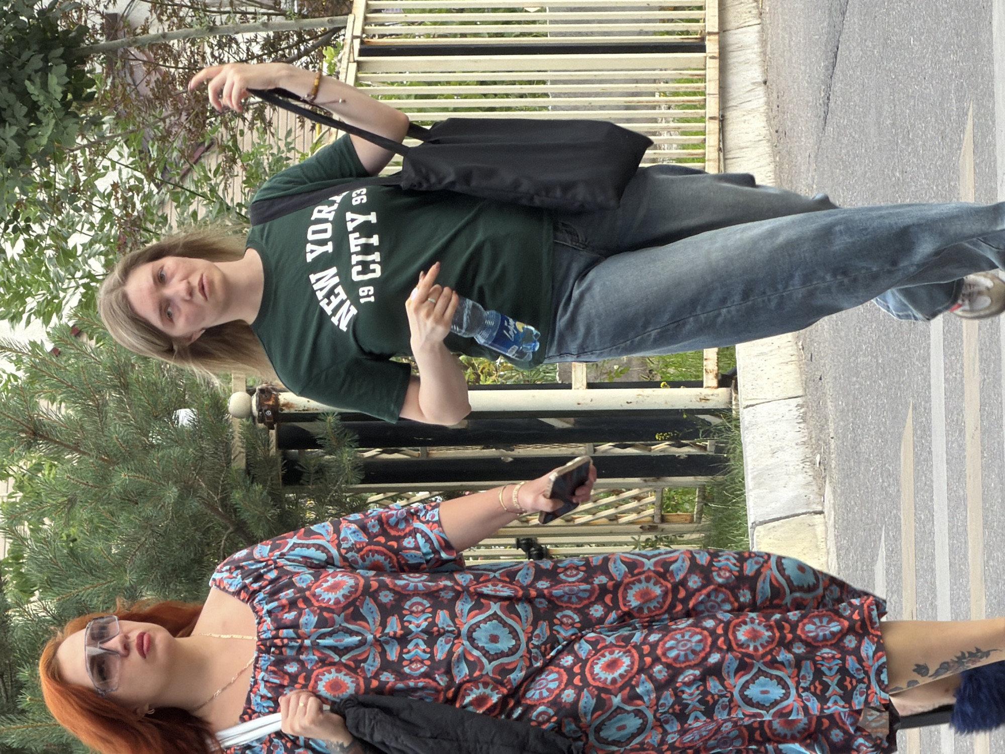 Two women are walking on a street. The woman on the left has red hair and is wearing sunglasses, a patterned dress, and carrying a black bag. The woman on the right has blonde hair and is wearing a green t-shirt with "NEW YORK CITY 93" printed on it, blue jeans, and carrying a black tote bag. She is also holding a water bottle and a cell phone. They are walking on a crosswalk in front of a metal fence and some greenery. The image appears to be taken in daylight.