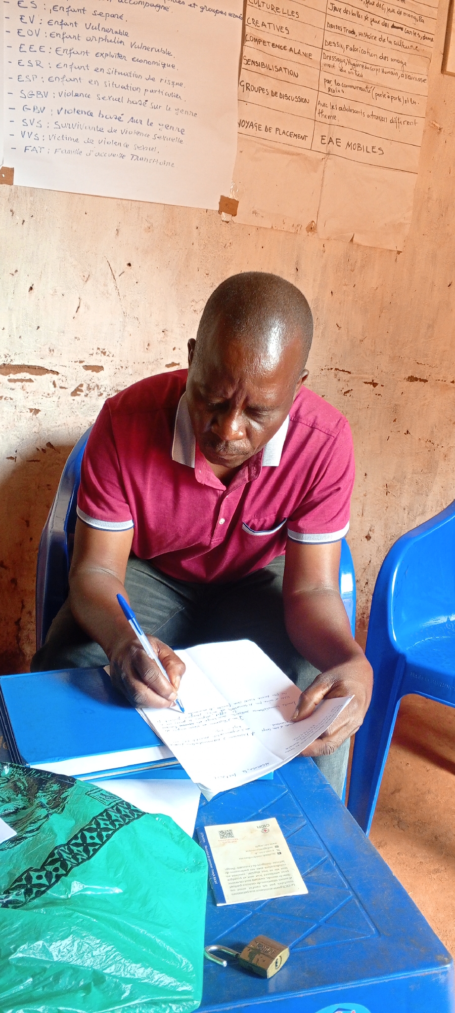 In Ngalula, Democratic Republic of the Congo, a man is seated on a blue plastic chair, intensely focused on writing in a notebook with a blue pen. He is wearing a maroon polo shirt with white trim and dark pants. On the table in front of him are a blue folder, a green bag with a dark pattern, a brochure from the ICRC (International Committee of the Red Cross), and a padlock. In the background, a wall is covered with two sheets of paper. The first sheet lists definitions, including 'ES: Enfant separe,' 'EV: Enfant Vulnerable,' 'EOV: Enfant orphelin Vulnerable,' and 'GBV: Violence bouse Aur le venire.' The second sheet lists activities such as 'CULTURELLES CREATIVES,' 'COMPETENCE ALANIE,' 'SENSIBILISATION,' and 'VOYAGE DE PLACEMENT EAE MOBILES.' The environment suggests an office or community education setting.