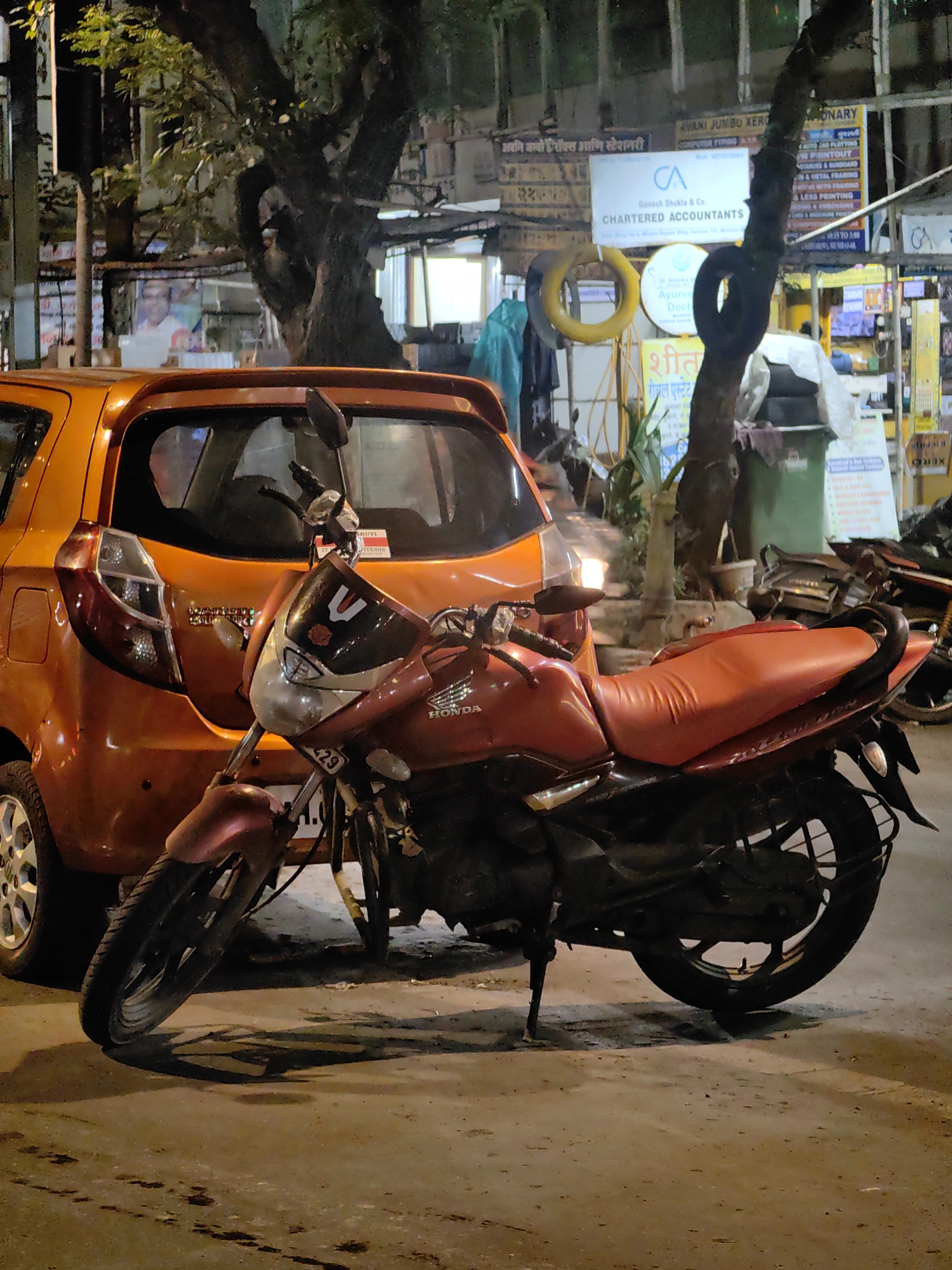 This is a photograph of a street scene in Mumbai, India, taken at night. A red motorcycle is parked in front of an orange car. The motorcycle has a black seat and chrome accents. The car appears to be a hatchback. In the background, there are buildings with signs, some of which are in Hindi. There are also trees and streetlights visible. The ground is a paved surface, possibly a road or sidewalk.