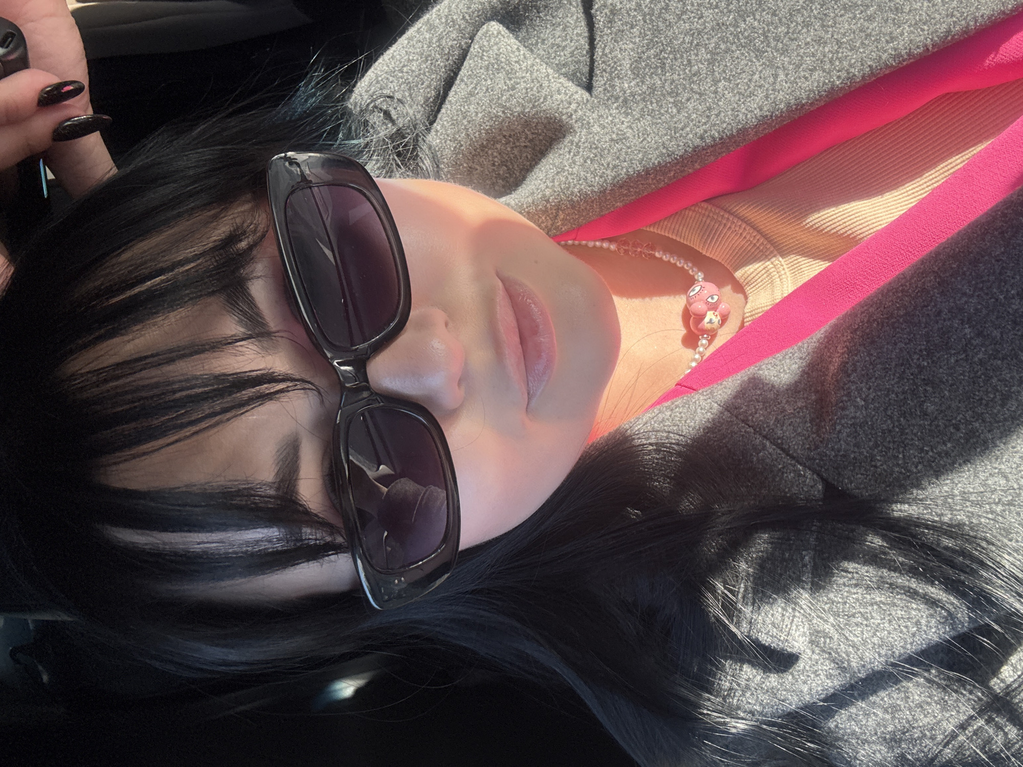 A young woman with black hair and sunglasses is pictured in a close-up shot. She is wearing a grey coat over a pink top, and a pearl necklace with a pink charm is visible. Her left hand, with long, dark fingernails, is holding something out of frame. The lighting suggests an outdoor setting with direct sunlight. The location is not specified in the image and cannot be determined to be the city of Chaikovsky, Russia.