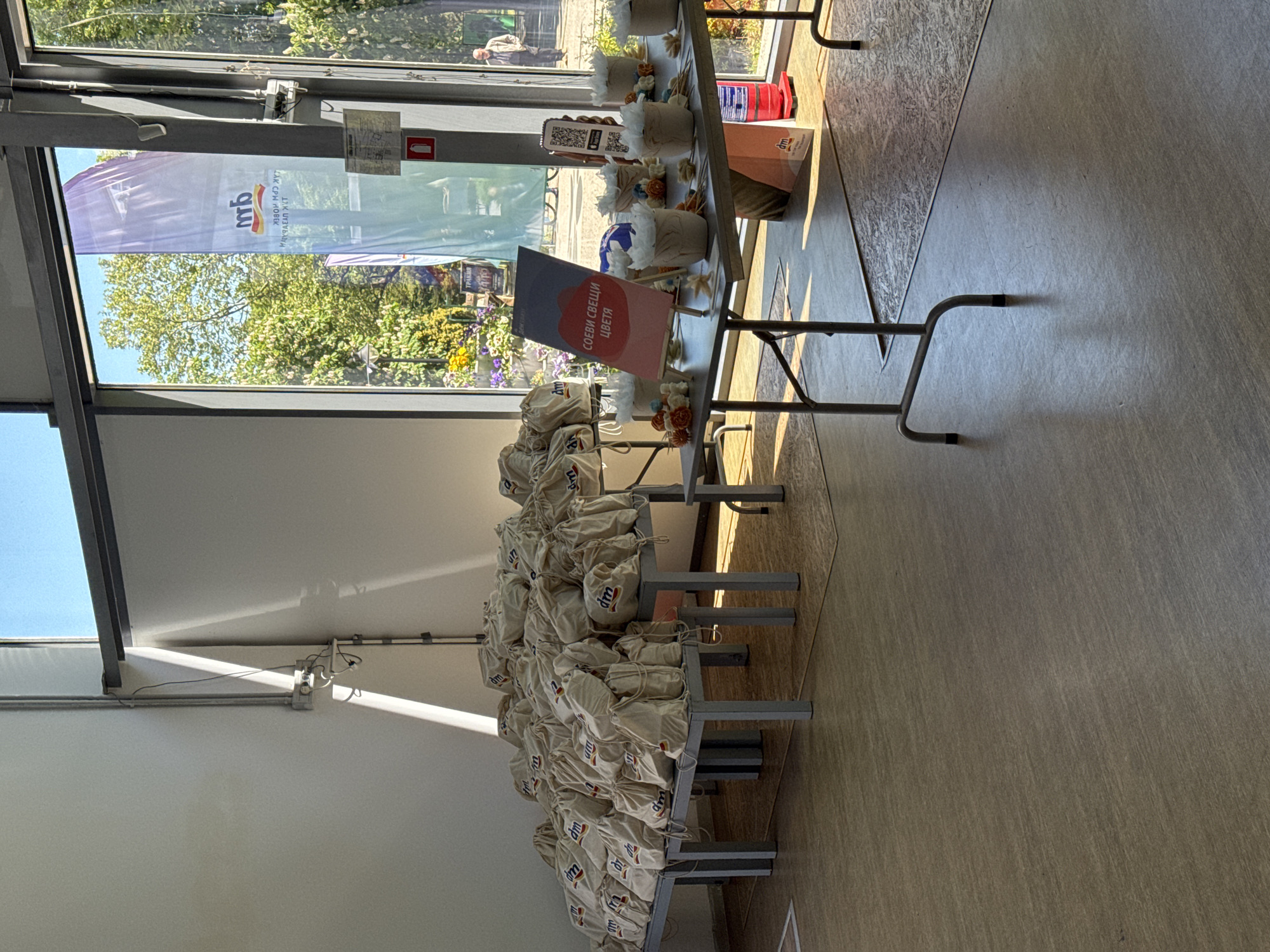 The image shows a promotional display in Burgas, Bulgaria. Tables are stacked with small canvas bags, each bearing the "dm" logo, indicating the drugstore chain. A sign in Bulgarian reads "Soy Candles, Flowers," suggesting the bags contain these items. The display is set up indoors, possibly in a community center or public space, as indicated by the large window showing a view of trees and an "mb" banner outside, and a fire extinguisher is visible behind the candle display. The light suggests it's daytime.
