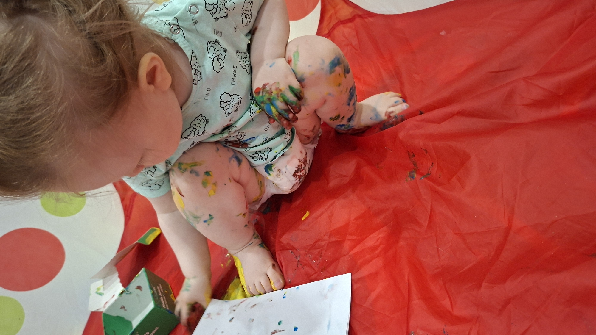 A toddler is sitting on a red plastic sheet with paint scattered around, likely engaging in a painting activity. The child is wearing a light blue patterned shirt and a diaper. Paint is visible on their hands, legs, and feet, suggesting they have been actively painting. The child is bending over towards a piece of paper and an open paint box. Part of a table with red polka dots can be seen in the background. There is no information to confirm location details.