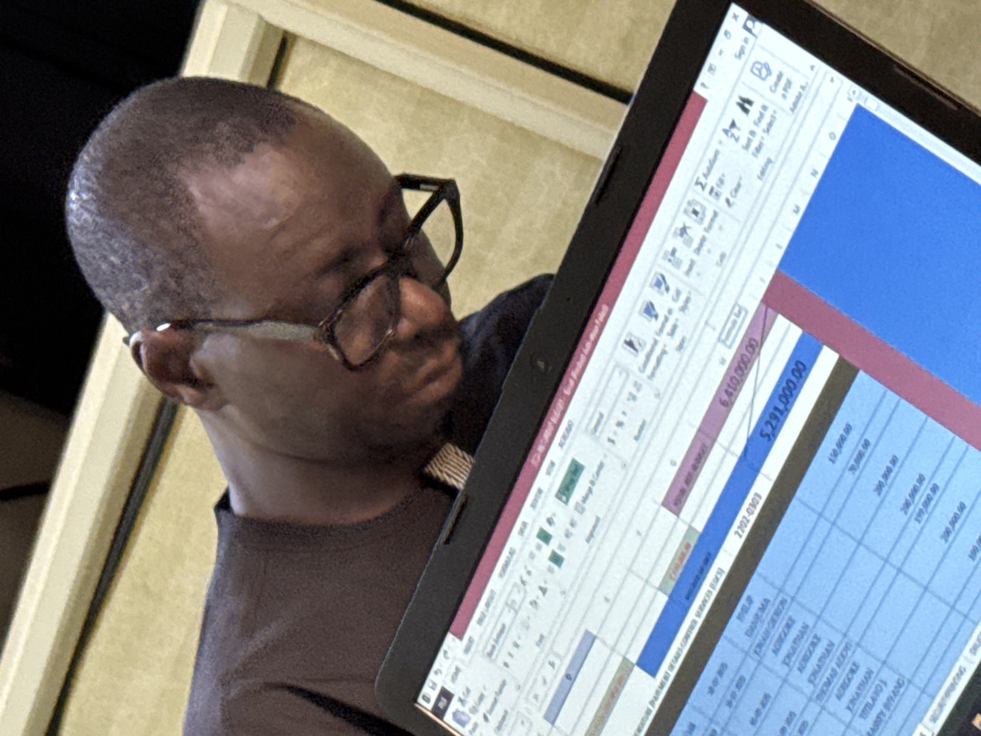 A dark-skinned man wearing glasses and a brown shirt is holding a tablet displaying a spreadsheet. The spreadsheet includes monetary amounts and names, including "Jonathan", "Adejoke", and "Mariity Biyang". The numbers listed are "6,410,000.00" and "5,291,000.00". I cannot determine the city of Abuja, Nigeria from the image.