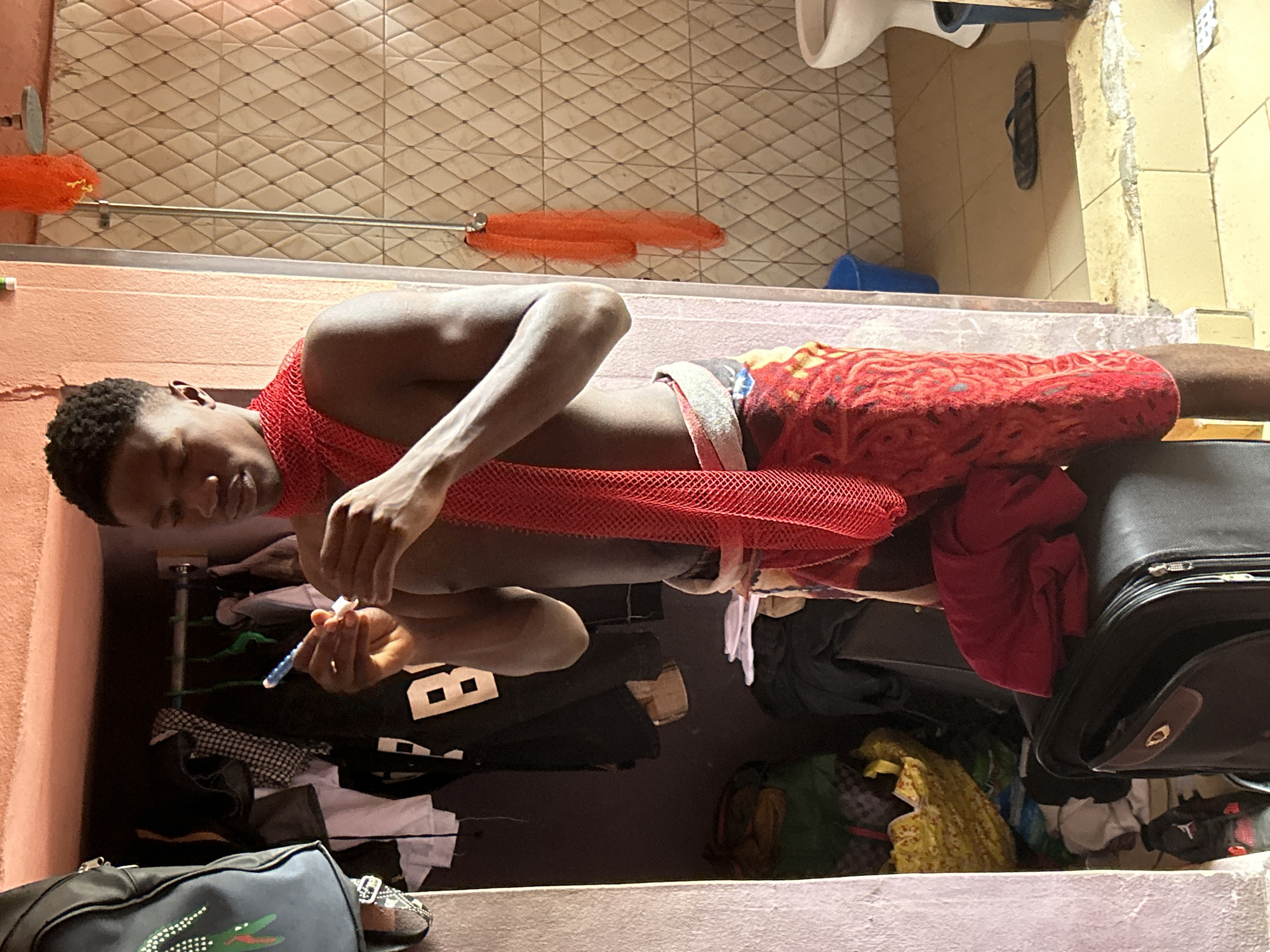 A young Black man is shown in a vertical orientation. He is seated on a surface and appears to be brushing his teeth with a blue toothbrush. He is wearing a red mesh garment around his neck and a red and white patterned cloth around his waist. His arms are extended forward, and his hands are focused on the activity.
The background suggests an indoor setting, possibly a bathroom or a cluttered room. A tiled wall is visible behind him, and a shower curtain rod with two orange netting-like objects is mounted on it. Various items are piled around him, including clothing, bags, and what appears to be a black suitcase. The overall impression is one of a lived-in, perhaps somewhat unkempt, space.
The lighting suggests it is daytime, but there are no clear indicators of time or weather. The man's expression is one of concentration. No visible text is discernible in the image. The location context of Ihiagwa, Nigeria, offers a cultural and geographical backdrop.
