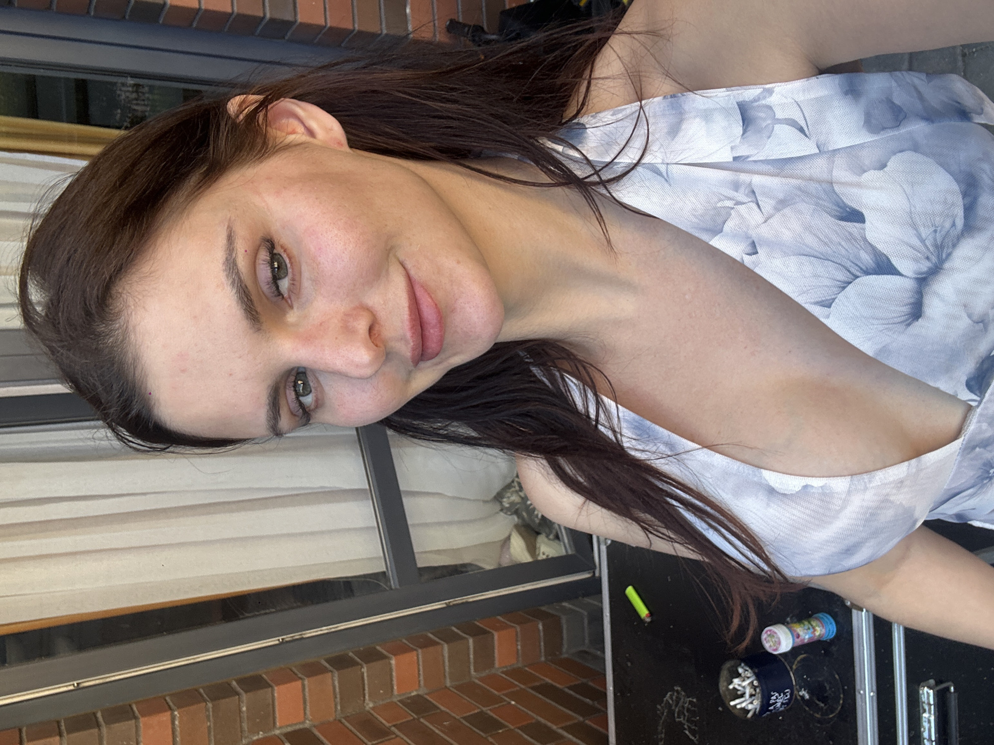 A young adult female with long, dark brown hair is captured in a close-up, slightly angled selfie outdoors. She is wearing a light-colored, floral-patterned halter dress with a deep neckline. Her gaze is directed towards the camera, and she has a subtle smile. In the background, there is a brick wall, a sliding glass door with vertical blinds, and a black surface with various objects including a metal container filled with cigarette butts, a lighter, and a small bottle with a colorful cap. The lighting suggests it is daytime.