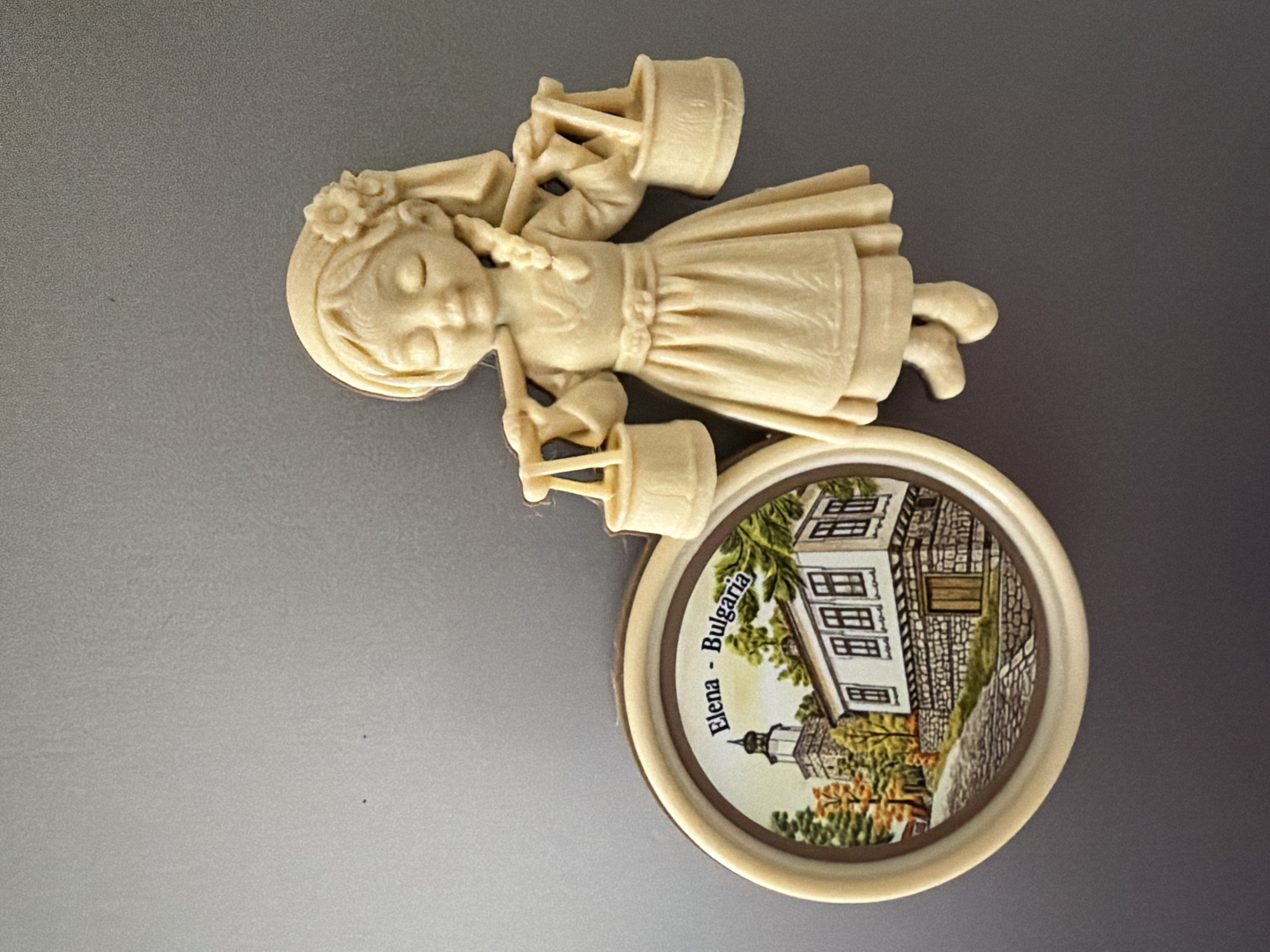 The image shows a souvenir magnet on a gray, textured surface. The magnet features two distinct parts: a circular scene and a figurine. The circular scene depicts a building with a clock tower, a stone wall, and trees with autumn foliage, along with the text "Elena - Bulgaria." To the right of the circular scene is a light-colored figurine of a young girl wearing a dress and a flower in her hair. She has her eyes closed and is carrying two buckets on a yoke over her shoulders. There are no people or actions depicted beyond the static presentation of the magnet. The environment is a plain, neutral gray surface, likely a refrigerator door. The location referenced on the magnet, Elena, is a town in the Stara Zagora Province of Bulgaria, not in the city of Pazardzhik.
