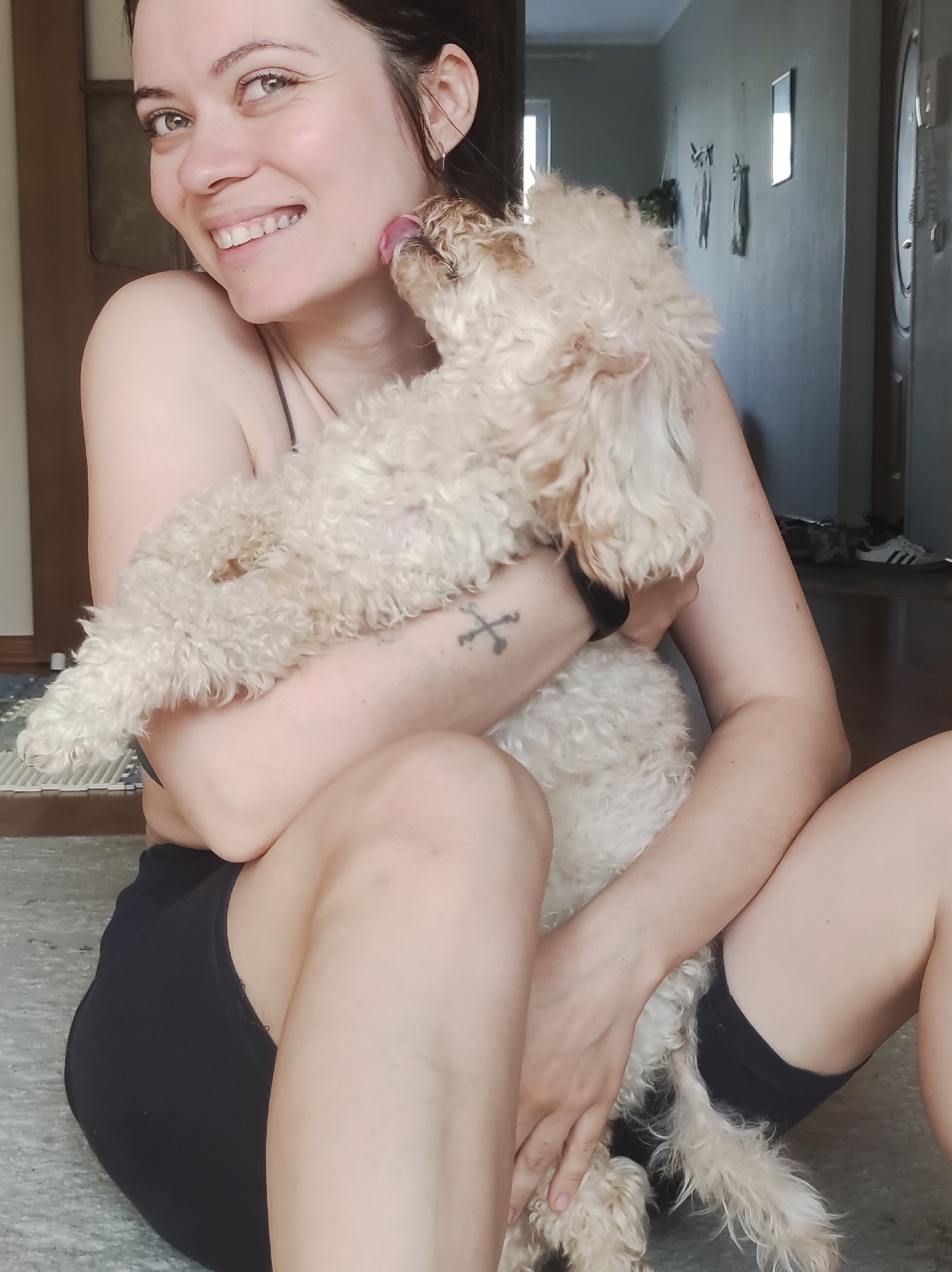A young woman with dark hair and bright green eyes sits on the floor, smiling widely and looking towards the camera. A small, fluffy, light-colored, curly-haired dog is held in her arms, enthusiastically licking her cheek. The woman wears a dark, strappy top and black shorts. A small tattoo resembling crossed lines is visible on her left forearm, along with a black wristband. The dog's light fur contrasts with her skin, and its tongue is visible as it licks her face.
The scene takes place indoors, in what appears to be a residence in Chaikovsky, Russia. The background features light-colored walls, a wooden doorframe, and a light-colored carpet or rug. On the far wall, a rectangular mirror and some indistinct decorative items are visible. Further in the background, a pair of white Adidas sneakers with black stripes can be seen on the floor, with "Adidas" visible on them. The natural, soft lighting suggests it is daytime. The overall mood is joyful and affectionate, highlighted by the woman's genuine smile and the dog's playful interaction.